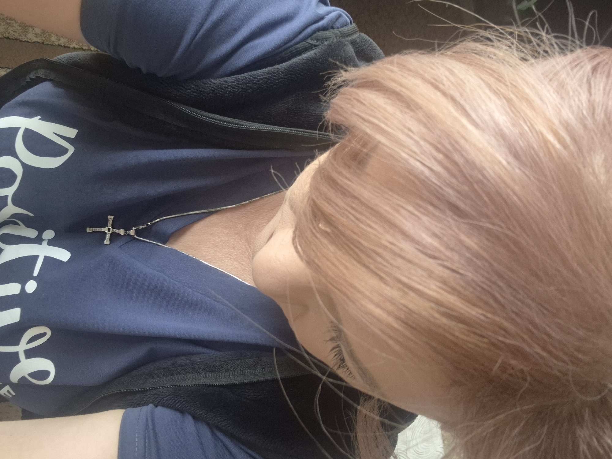 The image shows a close-up, overhead view of a person's head and upper body. The person has light pink or peachy colored hair that is styled in bangs. They are wearing a navy blue v-neck shirt with a fuzzy black vest or jacket over it. A silver chain with a cross pendant is visible around the person's neck. The text "Positive" in white, cursive lettering is visible on the blue shirt. The background is out of focus and appears to be indoors, with a hint of patterned flooring. The scene is likely in Vanadzor, Armenia, based on the provided context.
