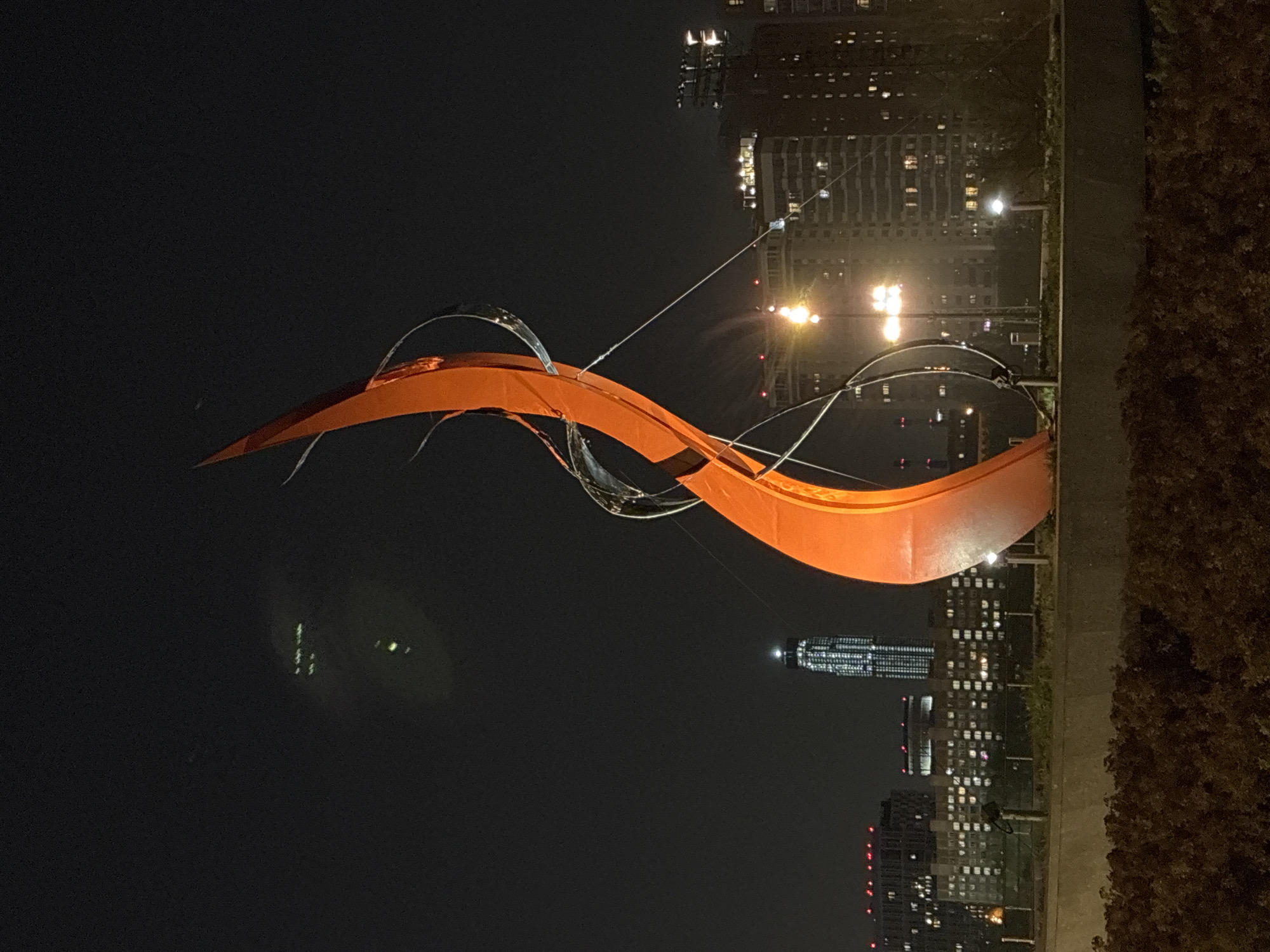 Here is a detailed description of the image:
The image shows an abstract orange sculpture with metal accents at night in an urban setting. The sculpture's shape is reminiscent of a ribbon or a wave. It is set against a dark night sky, with city buildings visible in the background. Lights from the buildings illuminate the sculpture, making it stand out prominently. The buildings are high-rise structures, suggesting a dense urban environment. There is also a strip of greenery with possible trees along the sculpture, a street lamp, and parts of a concrete ground, all on the right side of the frame.