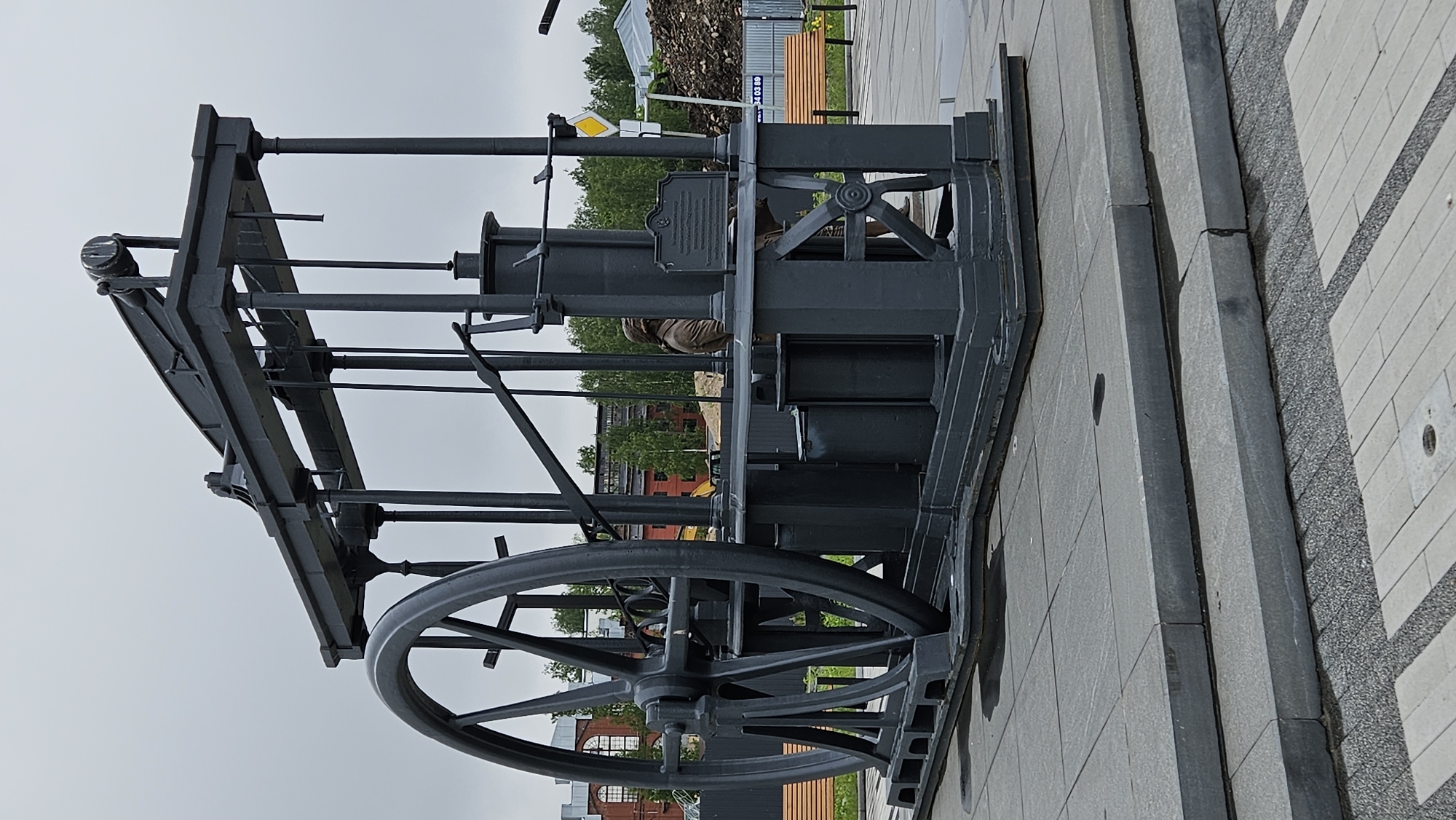 The image depicts a large, stationary, black metal machine with a prominent wheel on a paved surface. The machine appears to be an older piece of industrial equipment, possibly a stationary steam engine or a similar device. It has a framework of bars and supports, with the wheel positioned at the front. The background includes trees, a red brick building, a yellow sign, a bench and a pile of rocks or dirt. The overall setting suggests an urban or historical park. The image lacks specifics for a precise identification of the location, but it is in the city of Unknown, Unknown.