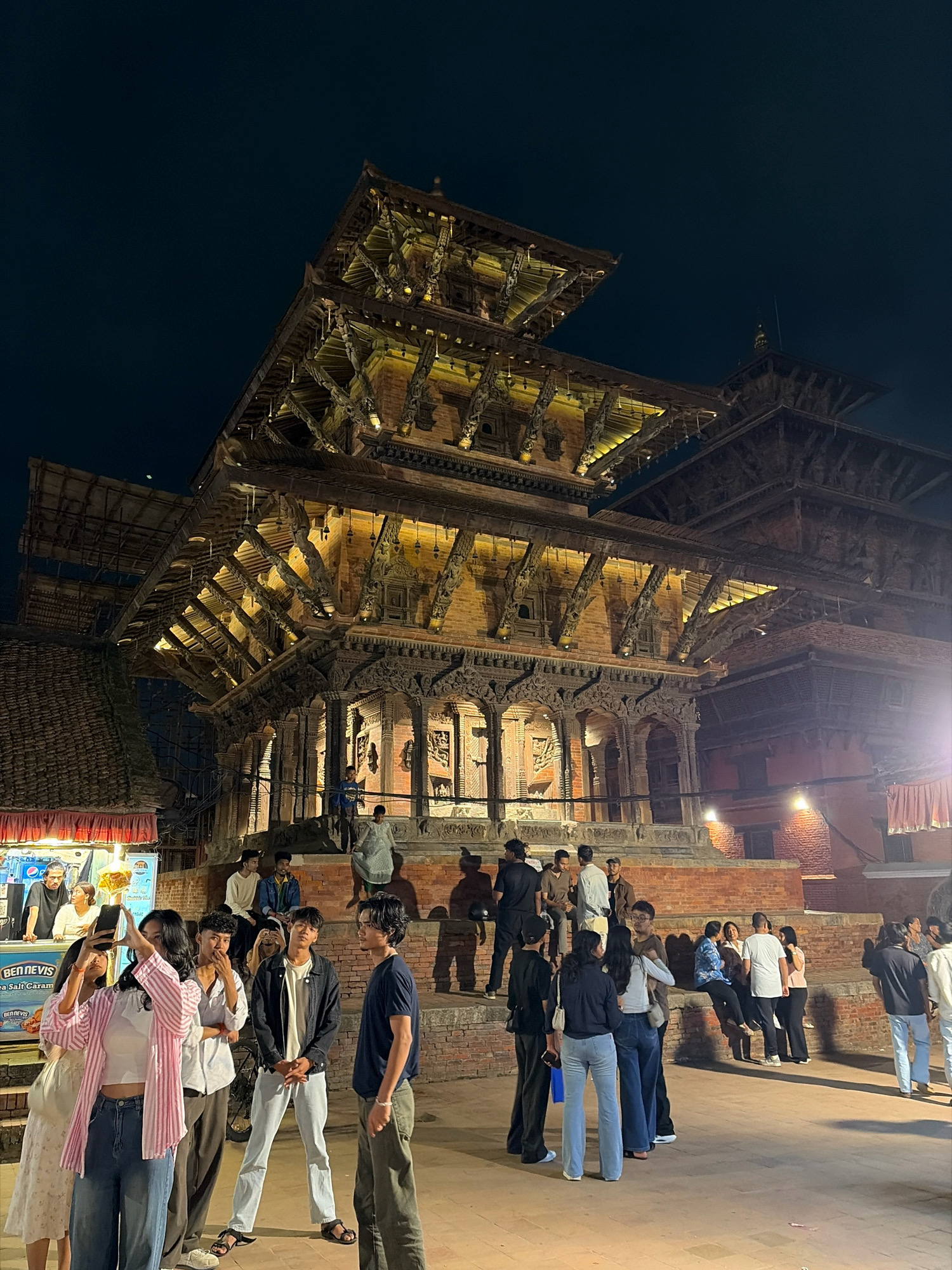 The image captures a nighttime scene in Patan Durbar Square, Lalitpur, Nepal, focusing on the intricately carved wooden architecture of the Taleju Temple. The temple, illuminated by warm yellow lights, dominates the frame with its multi-tiered pagoda-style roof and detailed latticework. Several people are gathered in the foreground and on the steps leading up to the temple, appearing to be tourists or locals enjoying the evening. Some are taking photos with their phones, while others are standing or sitting, engaged in conversation. A food stall with red and white striped awnings is visible to the left. The dark night sky provides a stark contrast to the illuminated temple, creating a dramatic and atmospheric setting. The ground is paved with stone, and shadows are cast by the people and the structures.