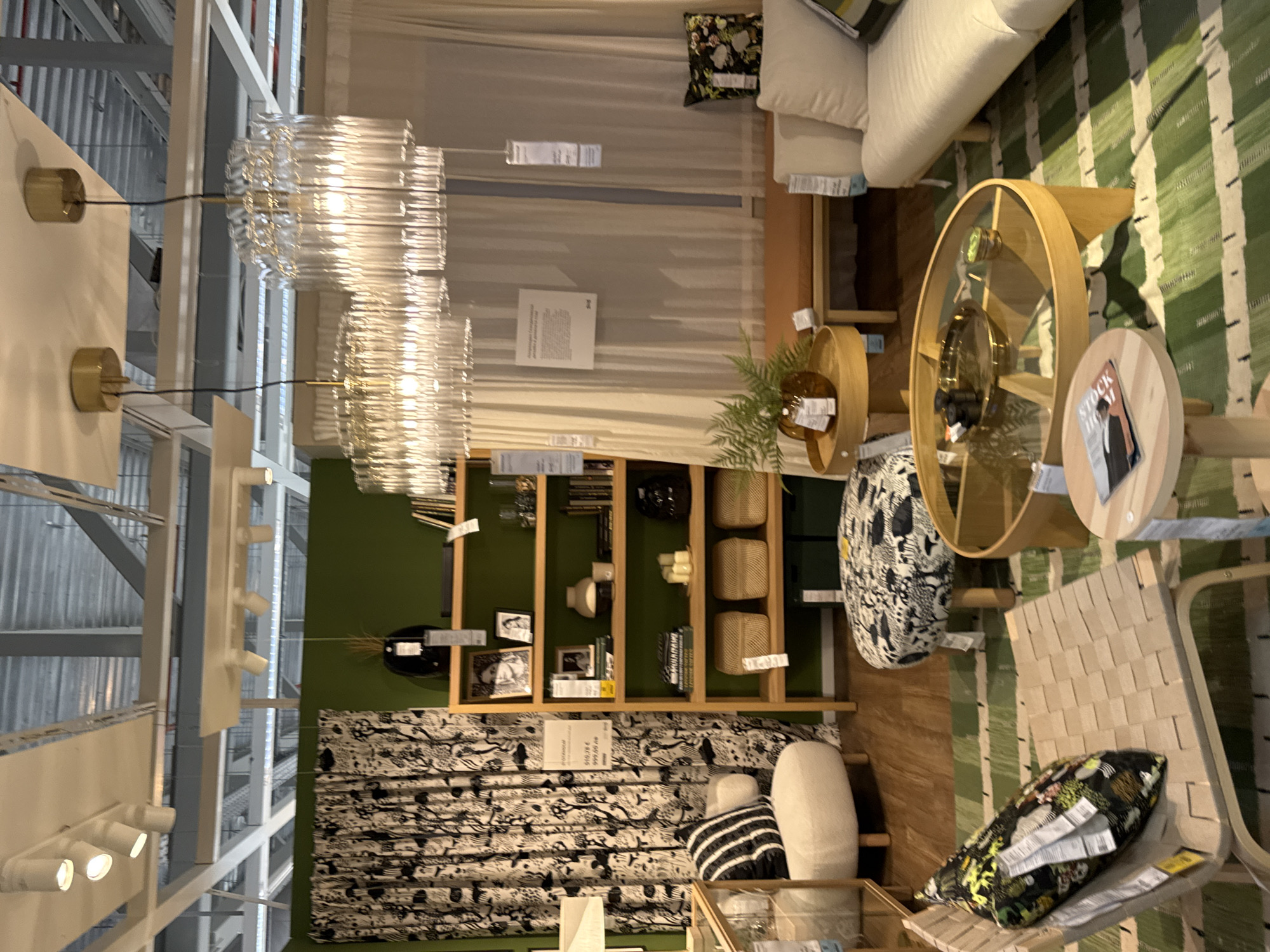 This is a medium shot, eye-level, indoor photograph of a modern living room or showroom display, likely in IKEA Sofia, Bulgaria. The scene features a variety of furniture and decor items arranged in a well-lit space.
In the foreground, a light-colored wooden lounge chair with a woven seat is positioned on a green and white striped rug. A decorative pillow with a black, green, and yellow botanical print rests on the chair. To the right, a round glass-topped coffee table with a wooden base sits on the rug. A small round side table is placed in front of the coffee table, holding a magazine.
Further back, an ottoman with a black and white patterned fabric cover stands on the rug. Behind it, a cream-colored sofa is visible, with a patterned pillow on its right end. A light wood bench is placed against a wall behind the sofa, with a tan cushion on top.
To the left of the scene, a plush, cream-colored armchair with a rounded back is situated near a patterned curtain with a black and white forest motif. A tall shelf unit with wooden shelves is against a dark green wall, displaying books, vases, framed pictures, and decorative boxes. A black vase with dried grass is on the top shelf.
Above the furniture, several modern light fixtures are suspended from the ceiling. Two elaborate glass chandeliers with multiple rods hang prominently, casting light on the scene. Other ceiling lights are also visible. The overall impression is one of a stylish and contemporary interior design.