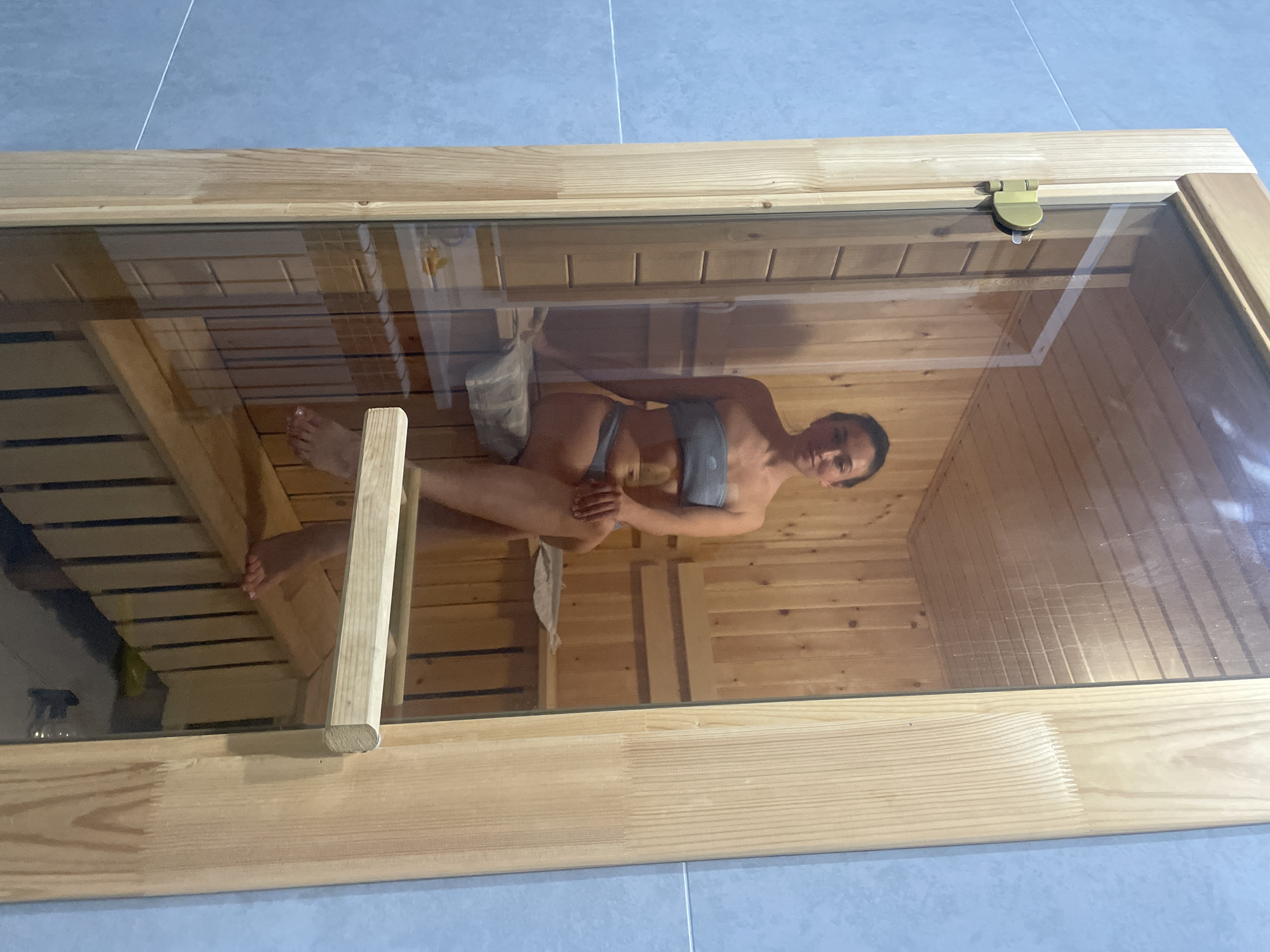 The image shows a woman in a sauna located in Эстачи, Russia. The woman is wearing a gray bikini or swimsuit and is sitting on a wooden bench with her legs crossed and arms resting on her knees. A white towel is placed next to her. The sauna has wooden walls and benches. The image is taken through a glass door or window with a wooden frame. The background shows a tiled floor in the room outside the sauna.