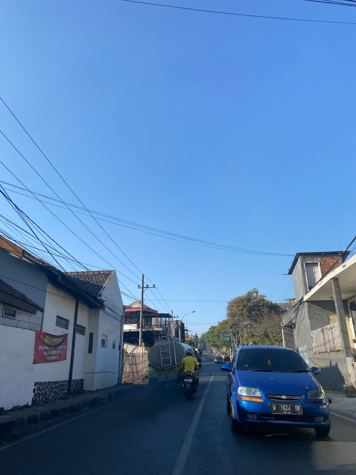 A street scene is captured under a clear blue sky. The perspective is from a moving vehicle, looking forward. On the left side of the street, there are several buildings with white walls and tiled roofs, and a red banner with Indonesian text is visible on one of the walls. Power lines crisscross overhead. On the right side of the street, there are more buildings, some with corrugated metal roofs and others with grey facades. A blue Chevrolet car with license plate "W 1624 BK" is in the foreground, facing the camera. Further down the street, a truck with a large tank is visible, and a person on a motorcycle wearing a yellow shirt is in front of it. The road is asphalt and appears to be in moderate condition. Trees with bare branches line parts of the street, suggesting a potentially cooler season. The overall impression is a bustling urban environment during daylight hours.