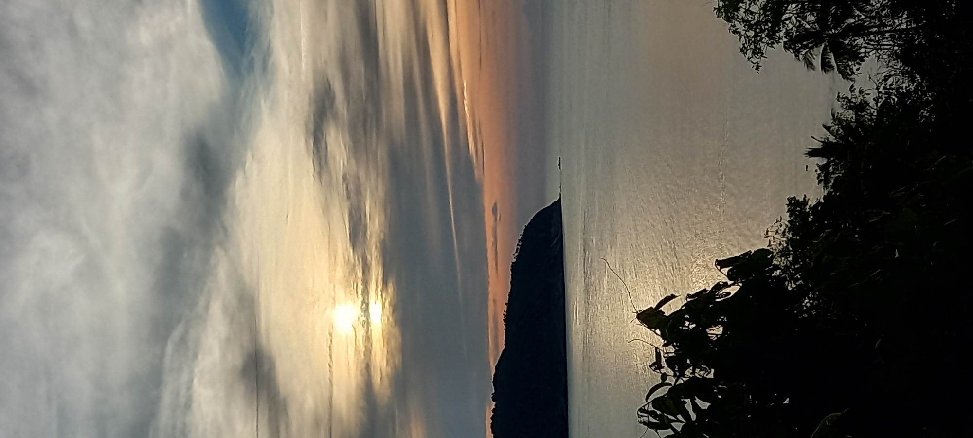 The image captures a sunset over the sea in Baan Chaloklum, Thailand. The sun is positioned low in the sky, partially obscured by clouds. Its light is reflected on the water's surface, creating a shimmering effect. A dark, mountainous landmass is visible between the sky and the sea. Silhouetted trees and foliage frame the right side of the image. The sky exhibits a gradient of colors, transitioning from blue to orange and yellow near the sun.