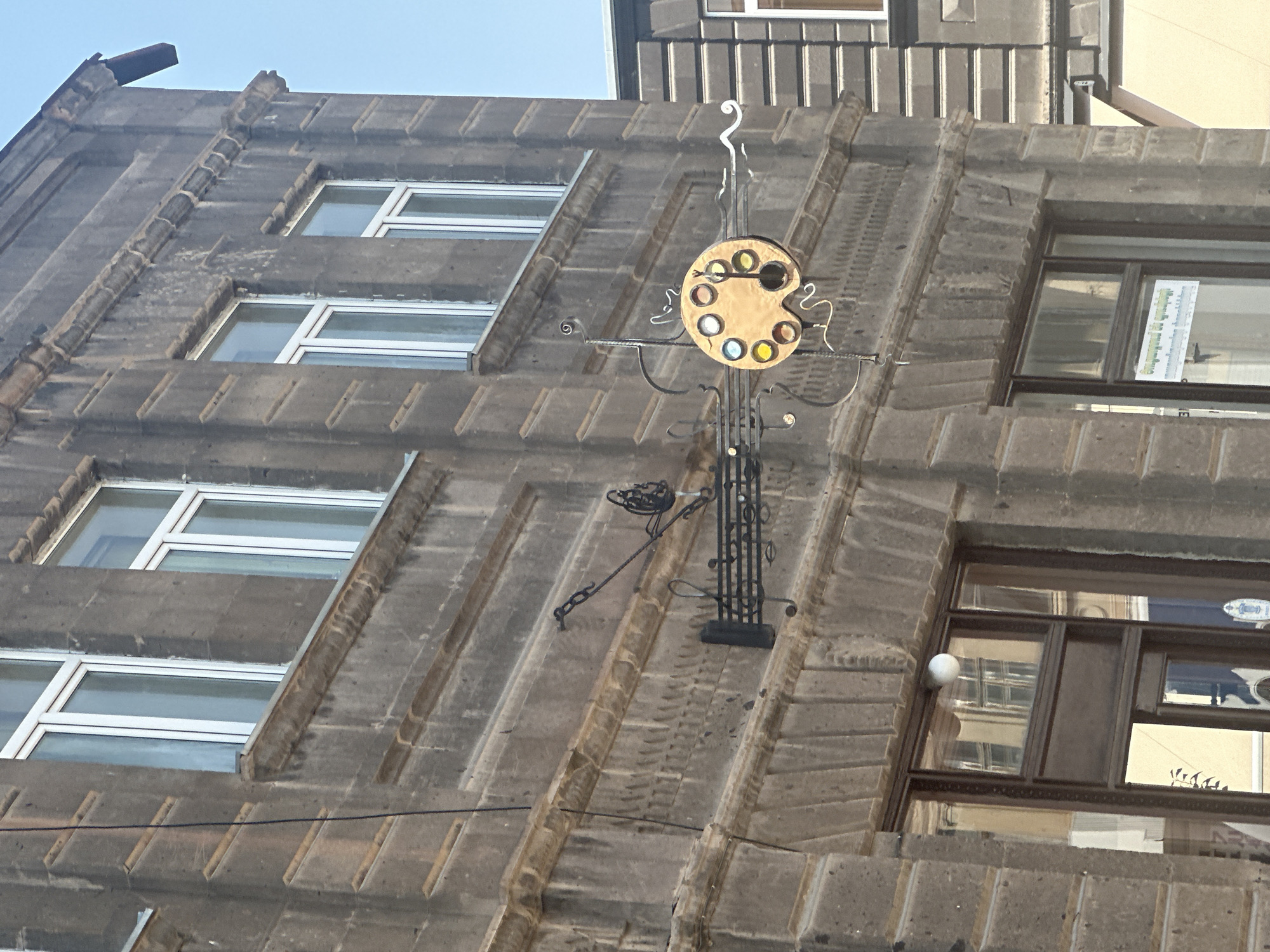 The image shows a street-level view of a building facade in Gyumri, Armenia. The building is constructed of dark grey stone with decorative detailing around the windows and cornices. There are several large windows with white frames visible on the upper floors.
A prominent feature in the center of the image is a metal sign hanging from the building. The sign is in the shape of an artist's palette, with a central cutout and several colored circles representing paint. The palette is attached to decorative metal scrollwork and music notes, suggesting a connection to art and music. Below the sign, a dark entrance with a glass door is visible, reflecting the light from across the street. A small, round light fixture is mounted above the door.
In the background, another building with a different architectural style and darker siding is partially visible. The sky above is clear and blue. The overall impression is one of an older, culturally rich urban environment.