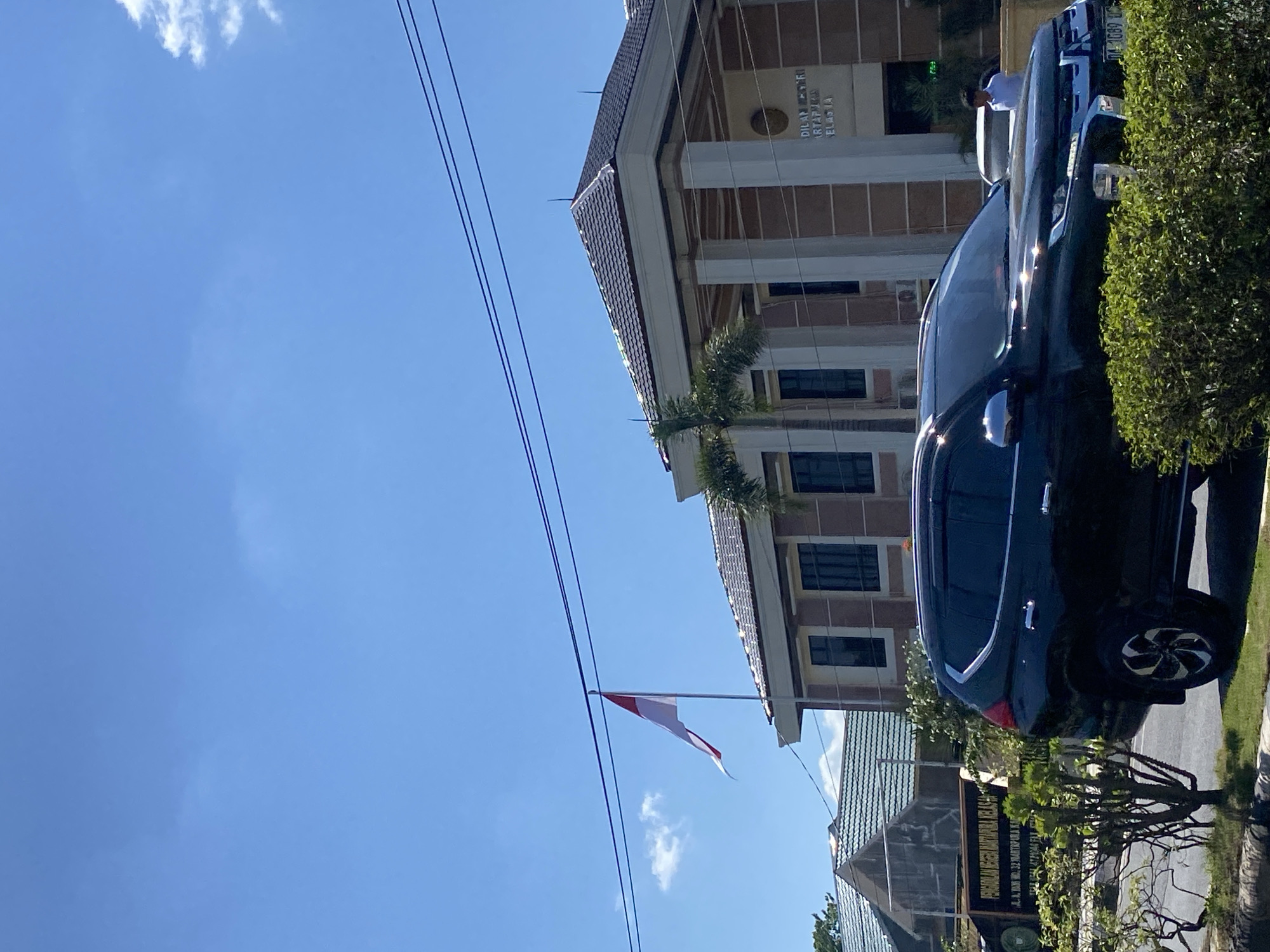 The image shows a clear, bright day in a city in Jawa, Indonesia. The foreground is dominated by the side and front of a black SUV parked on the side of a road, with lush green bushes surrounding it and a section of manicured lawn. Behind the SUV, a building with a tiled roof and cream-colored columns stands prominently. Above the building, an Indonesian flag, red and white, flutters on a flagpole against a bright blue sky with a few scattered white clouds. To the left of the main building, another building with a similar tiled roof is visible, and a sign with Indonesian text is mounted on a fence. In the background, another building and some trees can be seen. A person in a white shirt is visible near the entrance of the main building. The overall scene suggests an official or government building located in a well-maintained urban environment.