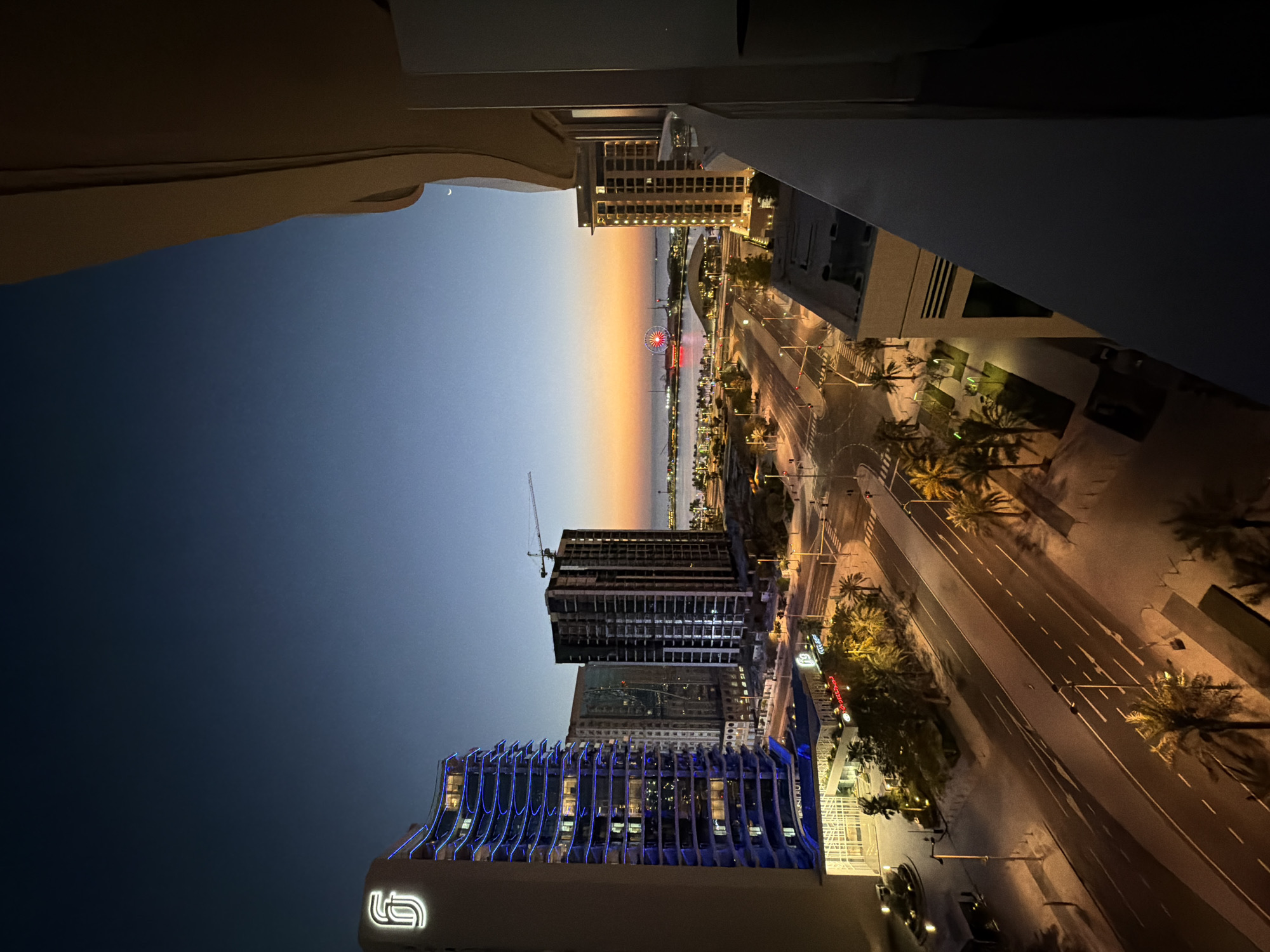 An aerial view captures a city skyline at dusk. The sky transitions from a deep blue at the top to a warm orange and pink gradient near the horizon. Several tall buildings dominate the scene, some illuminated with blue and white lights. A large Ferris wheel is visible in the distance on the waterfront, glowing red. A construction crane is perched atop one of the unfinished buildings. Palm trees line the streets and sidewalks, casting long shadows in the artificial light. The street below is paved and has white lane markings. A balcony railing in the foreground partially obstructs the view from the right side. The overall atmosphere is tranquil, with the city lights beginning to twinkle as daylight fades.