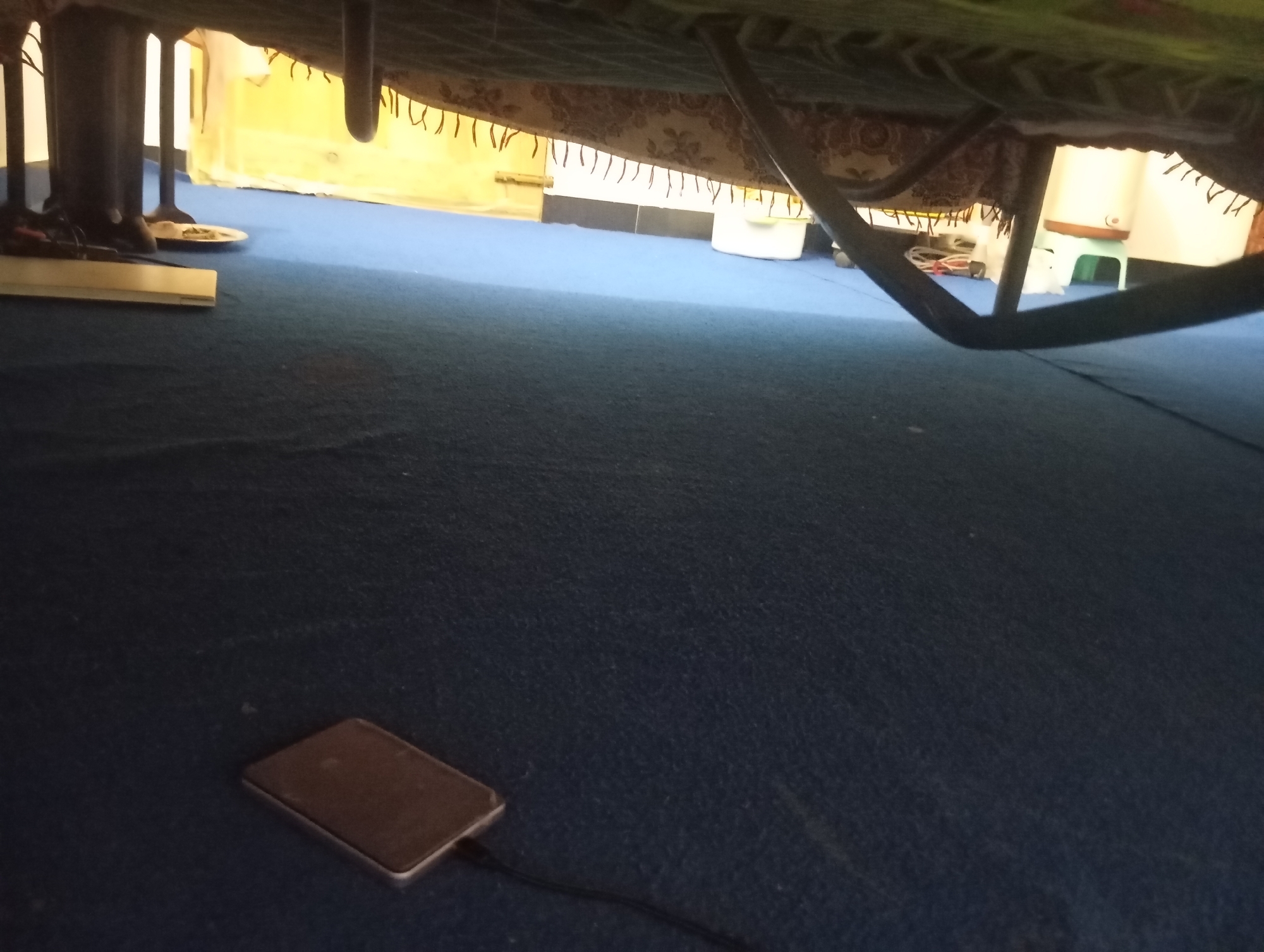 The image depicts the underside of a bed frame with a patterned fabric skirt hanging from it. The bed frame is supported by dark, cylindrical legs. In the foreground, a dark blue carpet covers the floor. A small, rectangular electronic device, possibly a power bank or portable charger, is situated on the carpet, connected to a black cable that extends out of frame. In the background, a light yellow wall is visible, with some white and brown objects, including what appears to be a small white stool. The overall scene suggests an indoor environment, possibly a bedroom, in a residential setting. There are no people visible in the image. The location of Lassan, Pakistan is not visually discernible from the provided image.