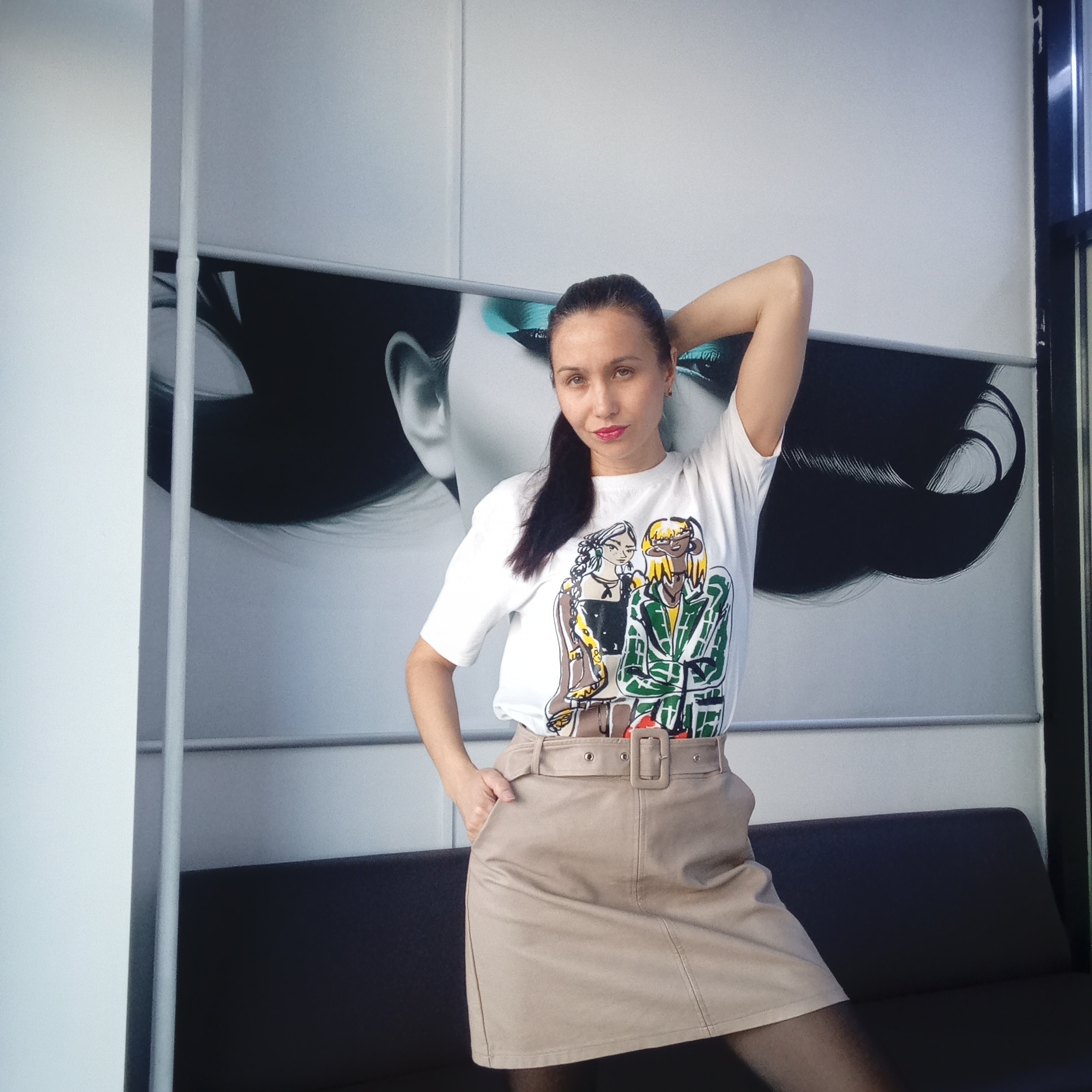 A woman is posing in a room, likely indoors, in Izhevsk, Russia. She has dark hair pulled back in a ponytail, fair skin, and is wearing red lipstick. Her left hand is in the pocket of a tan skirt with a matching belt. Her right arm is raised above her head, bent at the elbow. She is wearing a white t-shirt with a colorful graphic of two figures on it. Behind her is a wall with a stylized black and white image of a woman's face and hair. A gray metal bar runs horizontally across the wall. Below her is a dark couch. A white wall is visible to the left, and a window is visible to the right.