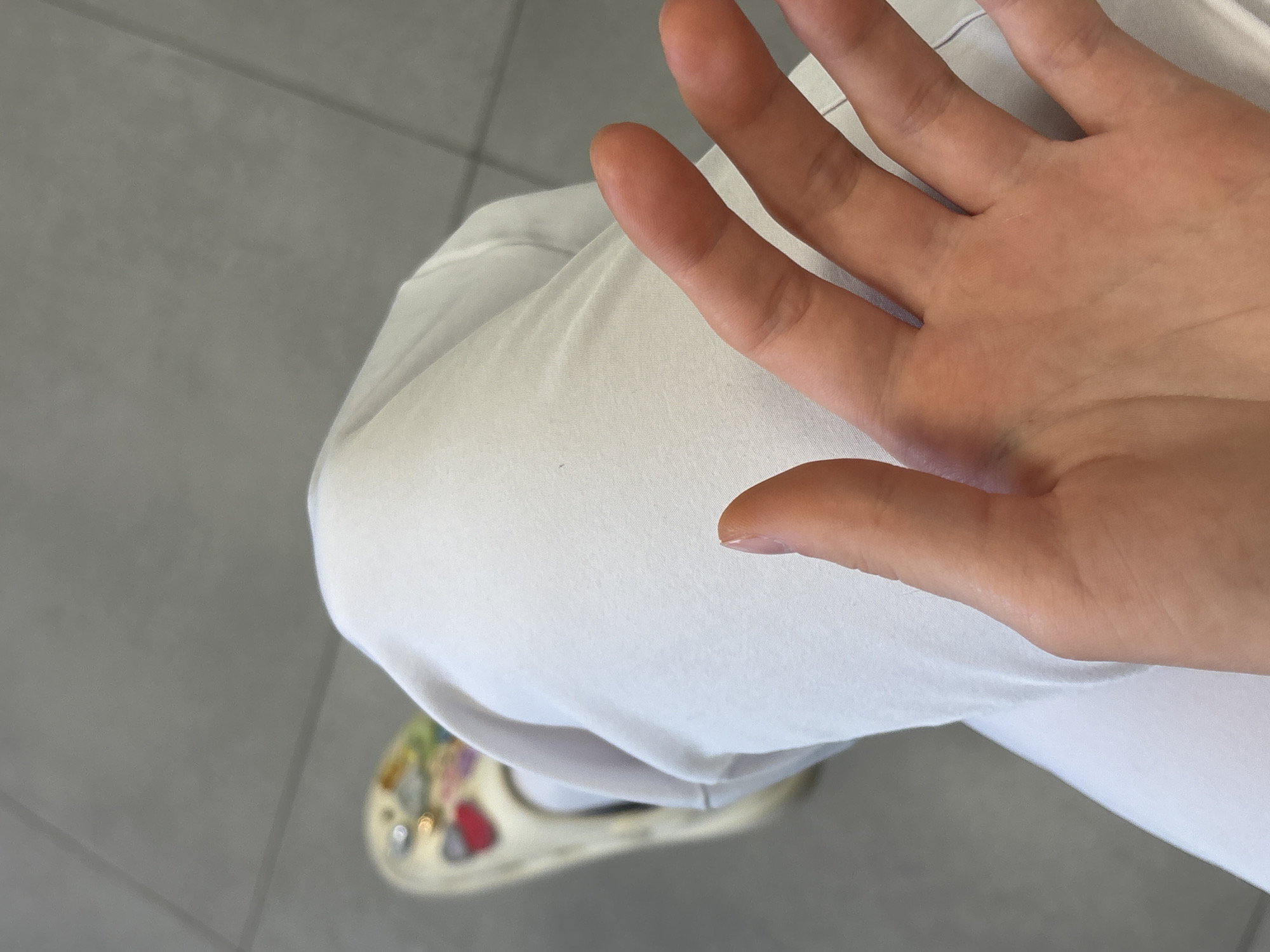 An overhead shot shows a right hand over a right thigh covered in white fabric, with a decorated slip-on shoe visible near the bottom left corner. The surface beneath the shoe appears to be gray tile. The location is not discernible from the image.