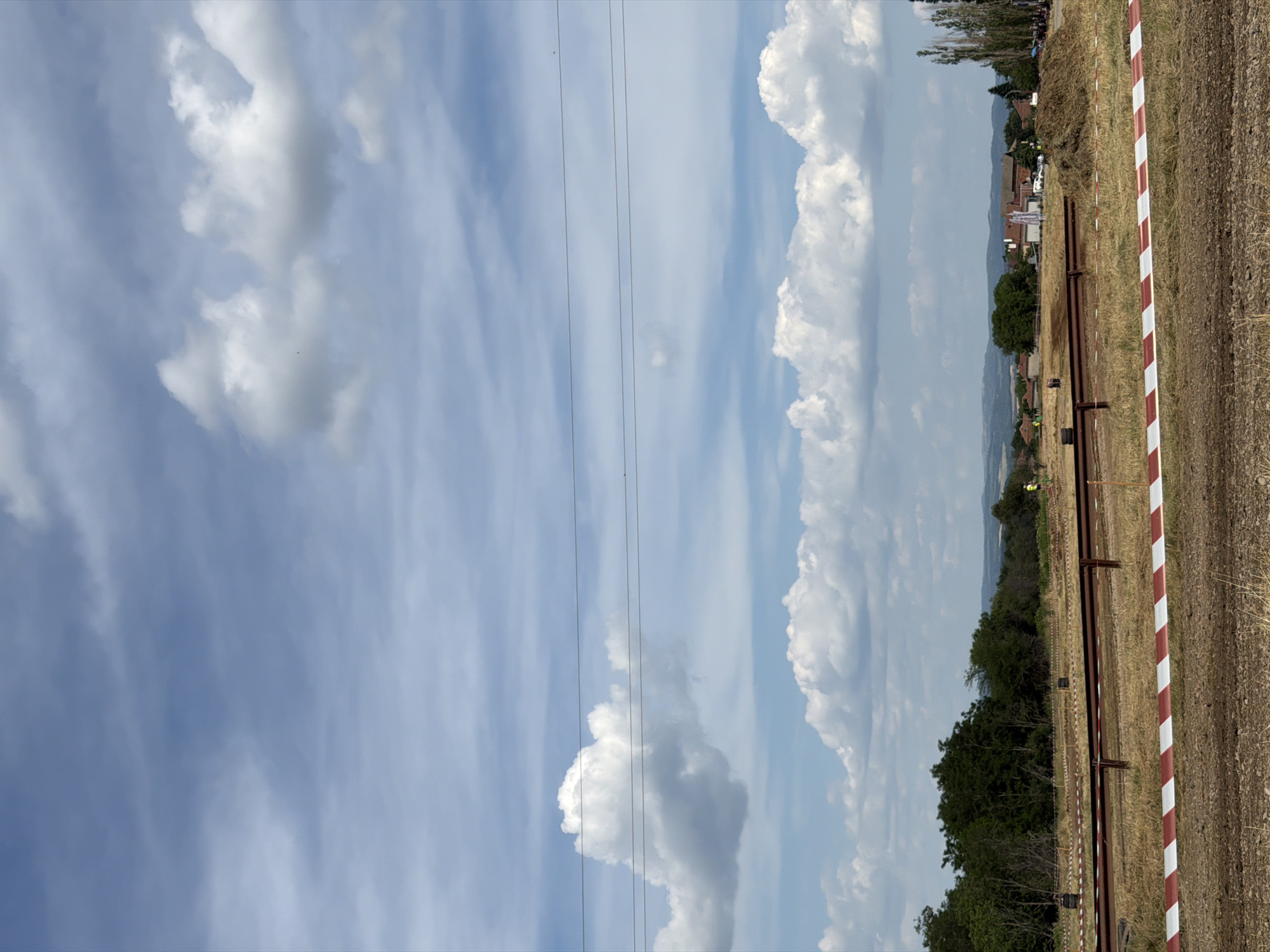 The image shows an outdoor scene with a cloudy sky. In the foreground, there is a dirt area with a red and white striped barrier tape. Beyond the tape, there is dry grass and a brown metal fence. In the middle ground, there are trees and some buildings in the distance, suggesting a rural or semi-rural setting. There are also several people visible, some standing near the fence and others further in the distance near the buildings. The overall impression is of an event or gathering taking place outdoors. The location is identified as Bahovitsa, Bulgaria.
