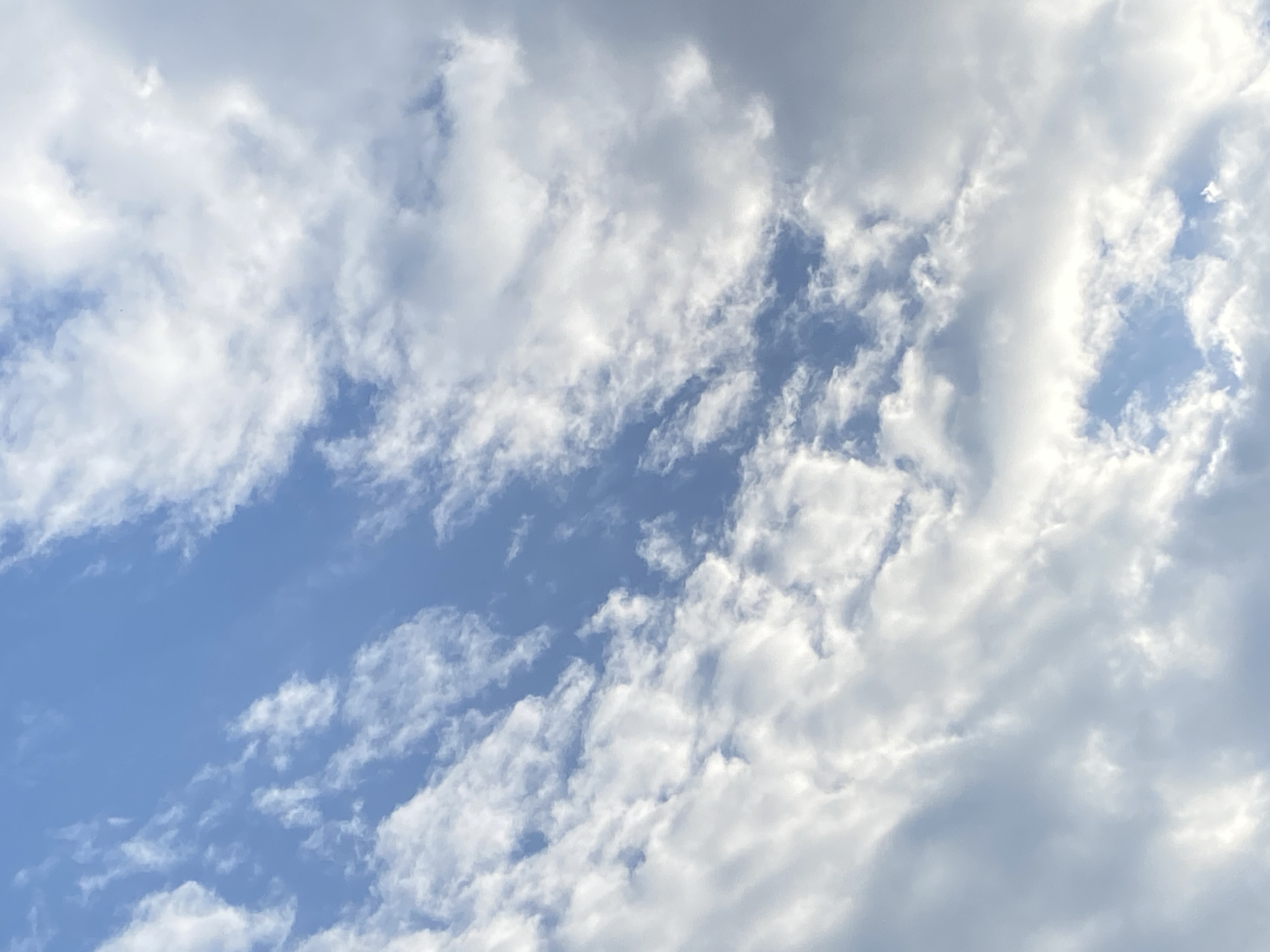 The image shows a daytime sky in Tinaquillo, Venezuela, partially covered with white and light gray clouds. There is no discernible urban environment or landscape visible besides the sky. The sky is a muted light blue in the areas not covered by clouds. No people, subjects, objects, actions, or interactions are visible.