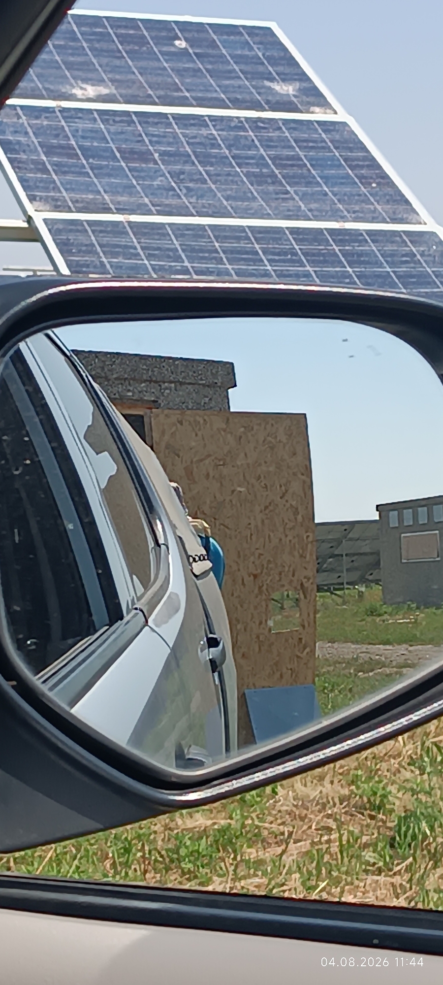 The image is a reflection in a car's side-view mirror, showing a scene in Starokozache, Ukraine. In the foreground of the reflection, a portion of a car's exterior, likely the driver's side door and window, is visible. Behind the car, a large wooden panel stands upright, obscuring part of the view. Further in the background, a building with several windows is visible, and beyond that, a field with dry grass and some green patches. A solar panel array is also present in the distance, angled upwards. The sky is clear and blue. The time stamp on the bottom right corner of the image indicates it was taken on April 8, 2026, at 11:44. There are no people visible in the reflection.