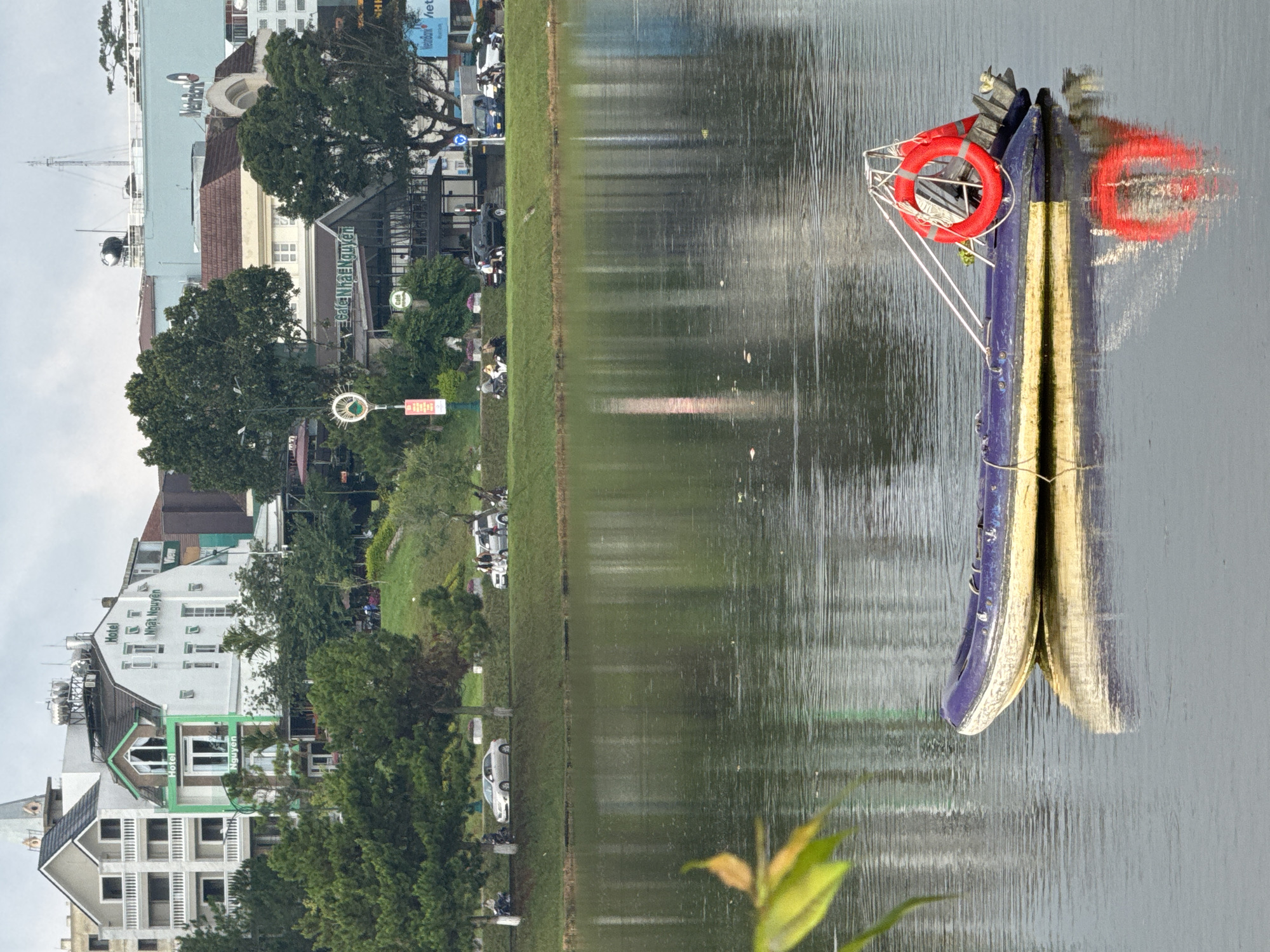 This image captures a serene view of Xuan Huong Lake in Da Lat, Vietnam, under an overcast sky. In the foreground, a small, purple and white inflatable boat with a red life preserver is anchored on the water. The boat features a small outboard motor and a metal frame. Reflected in the calm water are the boat and the surrounding trees and buildings. Across the lake, a tree-lined embankment separates the water from the city, where a variety of buildings are visible. These include multi-story hotels and shops, some with distinctive architecture and signage, such as "Hotel Nhat Nguyen" and "Cafe Nha Nguyen". A few cars and motorbikes are present on the road bordering the lake, and several individuals are visible walking or riding. The overall atmosphere is tranquil, with the lush greenery of the trees contrasting with the urban backdrop.