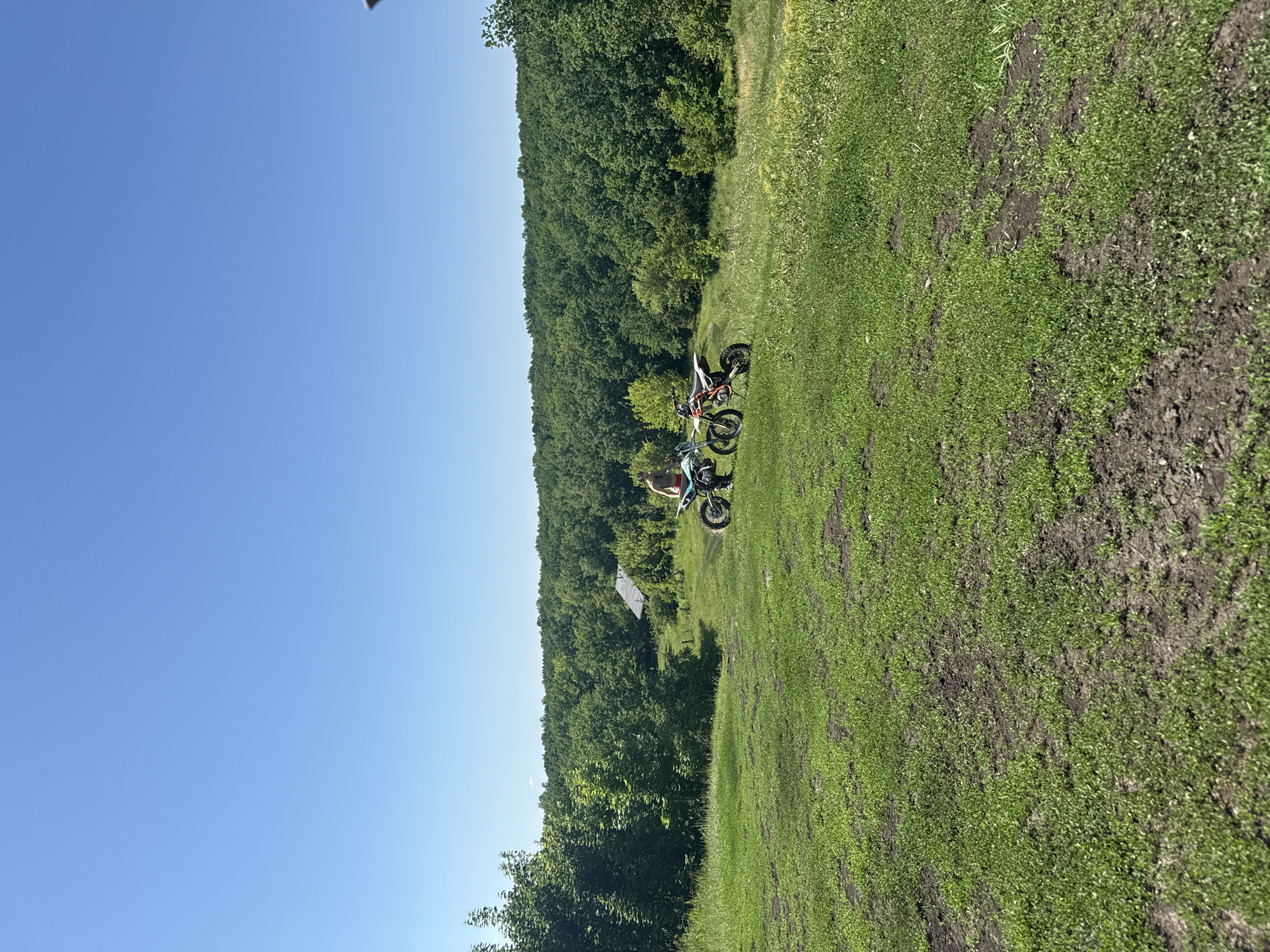 On a sunny day with a clear blue sky, a young person is standing next to two dirt bikes on a grassy hillside. The hillside slopes down towards a wooded area with a small house visible amongst the trees. The person is wearing red shorts and a light-colored top. One dirt bike is white and black, and the other is orange and white. The foreground of the image is dominated by the green grass of the hillside, with patches of bare earth. The background features a dense forest. The scene appears to be a rural or semi-rural location.