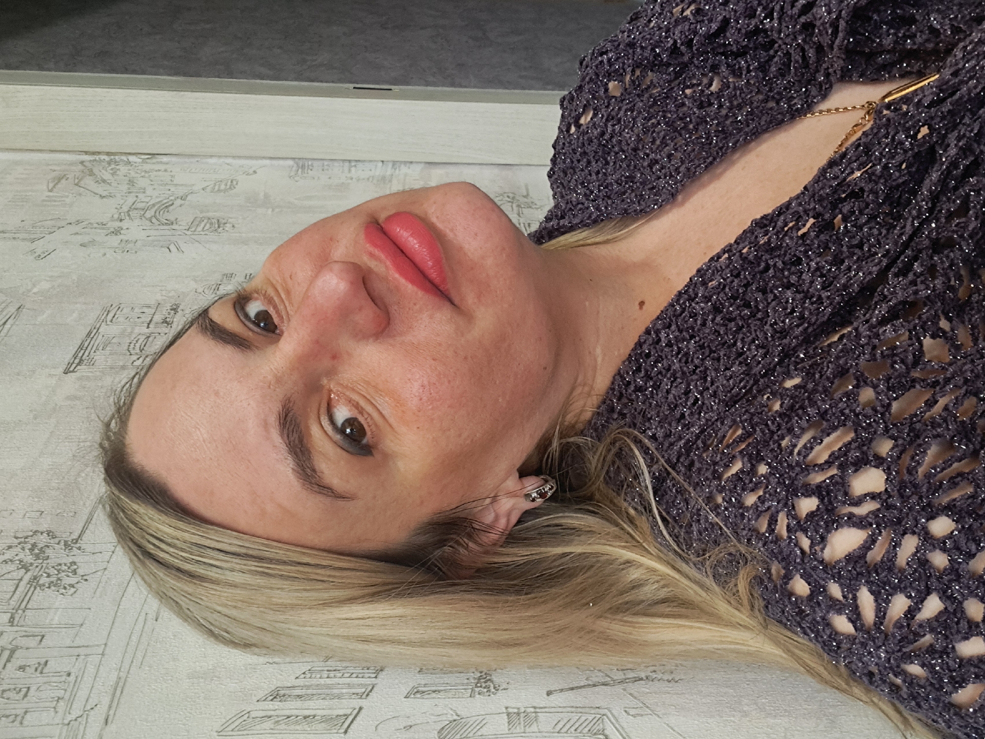 A woman with fair skin, blonde hair, and brown eyes is shown in a medium close-up shot. She is wearing a dark, patterned top with a hint of sparkle and a gold chain. The background appears to be a wall with a light-colored, patterned wallpaper. The photograph does not contain information to suggest the location is Elektrostal, Russia. The image does not show specific actions or interactions. The woman is looking directly at the camera with a slight smirk.
