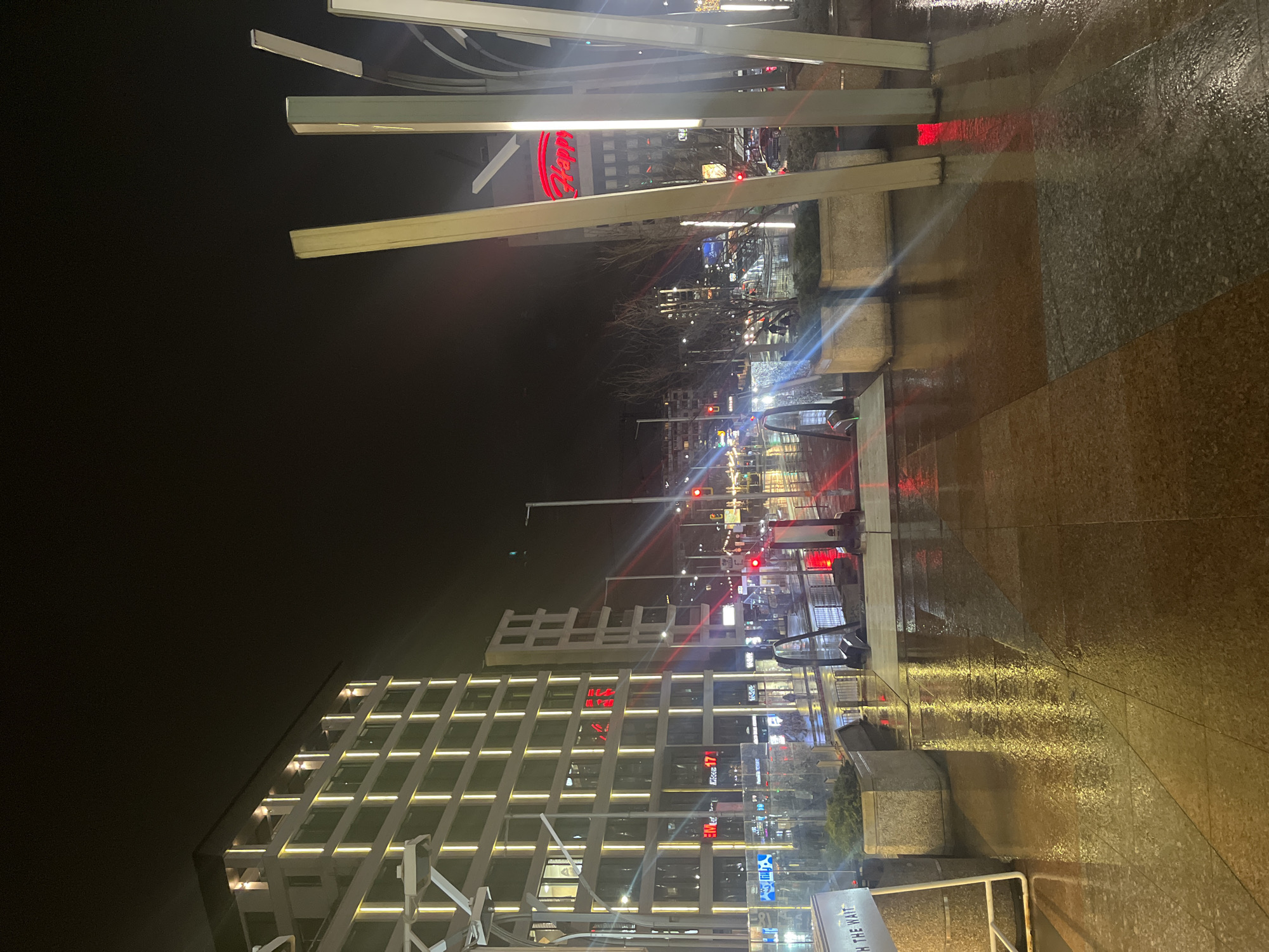 The image shows a nighttime urban scene in Sofia, Bulgaria. The foreground features a paved pedestrian area, likely a sidewalk or plaza, with wet pavement reflecting the city lights. Architectural elements are visible, including modern light fixtures and a building with a grid-like facade illuminated by white lights. Streetlights and traffic signals create streaks of red and white light. In the middle ground, traffic moves along a street, with several lit signs and storefronts. In the background there is a red lit sign that says 'happy'. The overall impression is a rainy or wet night in a contemporary urban environment.