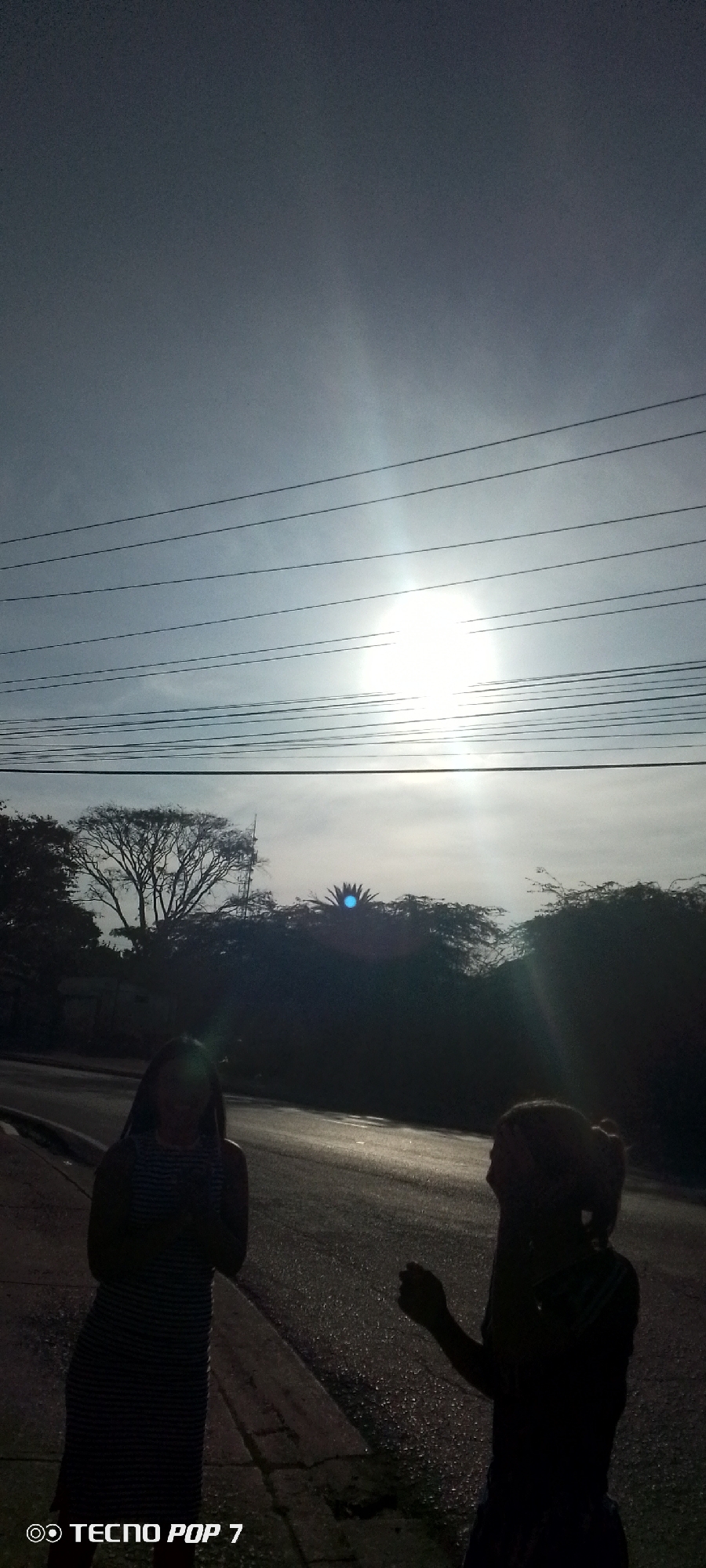 The image depicts two individuals silhouetted against a brightly lit sky in what appears to be the city of Altagracia, Venezuela. The setting seems to be an asphalt road or sidewalk. The sky is partly cloudy, with the sun obscured behind a layer of haze or clouds, creating a strong glare. Several power lines stretch across the upper portion of the frame. A line of trees and vegetation forms a darker, blurry backdrop behind the subjects. The silhouetted figures stand near the roadside, but their faces are not clearly visible due to the strong backlighting. The image includes a watermark at the bottom left corner, indicating the device "Tecno Pop 7."