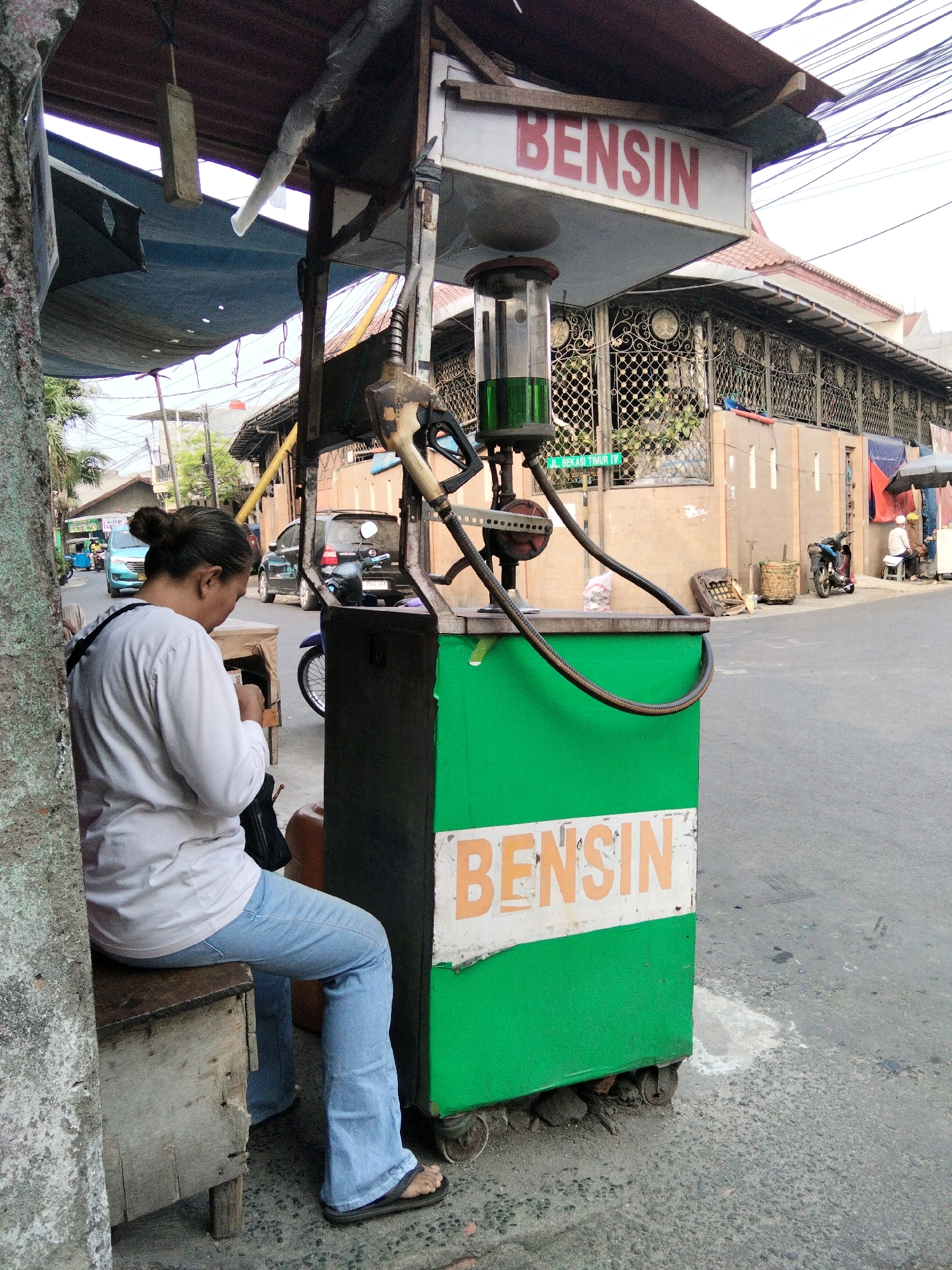 In East Jakarta, Indonesia, a woman sits on a wooden crate next to a roadside gasoline pump. The pump, painted green and white, has a sign that reads "BENSIN" in red letters. A clear glass cylinder on top of the pump contains green liquid, likely gasoline. A fuel hose and nozzle hang from the pump structure. The woman, wearing a white long-sleeved shirt and blue jeans, faces away from the camera, looking down at something in her hands. She has her hair tied up in a bun and wears black flip-flops. Behind the pump, a street runs perpendicular to the camera's view, with buildings and other structures lining its sides. A blue car and a motorcycle are visible on the street. The overall scene depicts a common sight in urban areas of Indonesia where small, informal gasoline stations are prevalent.