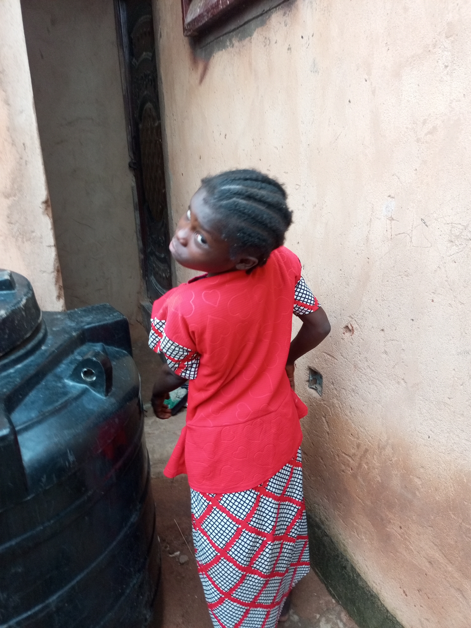 A young Black girl with braided hair is standing in an alleyway, viewed from behind and to her left, her head turned back towards the camera. She is wearing a red shirt with a heart pattern and a long skirt with a black and white checkered pattern. To her left is a large, black plastic water tank. The background consists of a textured, light brown wall and a dark, recessed doorway. The ground is dirt. The scene appears to be in an outdoor urban setting in Gombe, Nigeria.