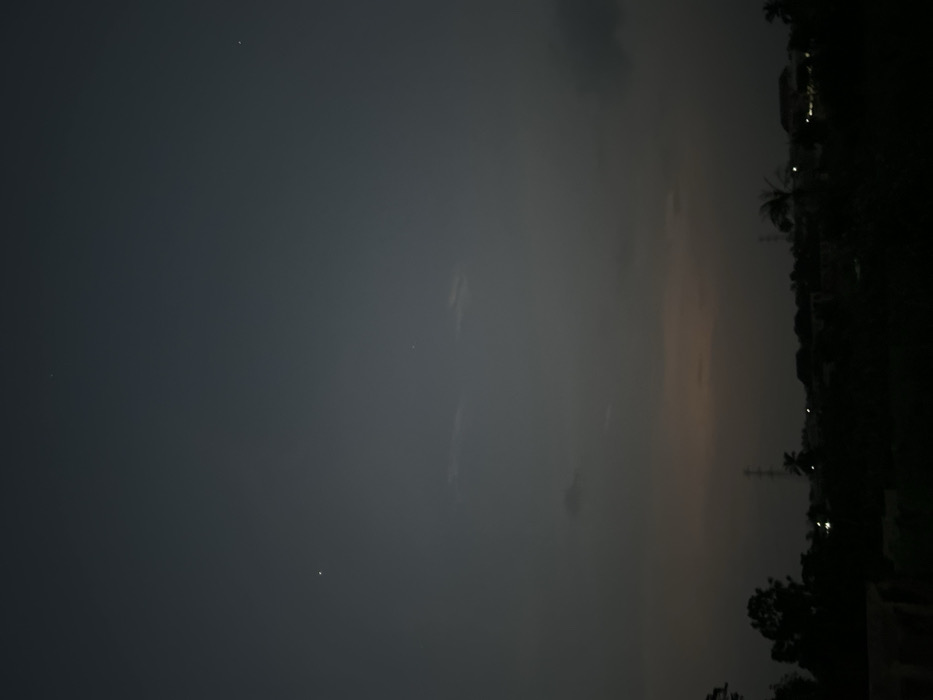 The image depicts a serene, low-light scene, likely captured during dusk or nighttime, dominated by a vast sky over a silhouetted landscape. The image is oriented vertically but will be described as if rotated to its natural horizontal view.
The upper two-thirds of the image are occupied by a dark, deep grey-blue sky, indicating very dim lighting conditions. Scattered across this dark expanse are several tiny, pinprick white lights, which could be stars, planets, or very distant artificial light sources. Subtle, wispy cloud formations are visible, particularly in the middle and right sections of the sky, appearing as slightly lighter grey patches against the darker background.
Towards the right side of the image, closer to the horizon line, there is a distinct and diffused glow. This glow exhibits a soft gradient, transitioning from a faint orange-pink hue near the bottom-right to lighter shades of grey further up, blending into the darker sky. This suggests either the last remnants of twilight or significant light pollution from an urban area.
Below this illuminated horizon, a dark, indistinct silhouette of a landscape stretches across the lower third of the image. This silhouette comprises various shapes, including dense foliage that appears to be trees, some with recognizable fronds suggestive of palm trees. Interspersed within this dark outline are several small, bright artificial lights, likely originating from buildings, streetlights, or other structures in the distance. Vertical structures, possibly utility poles or towers, are also discernible against the brighter sky.
The overall impression is one of quiet contemplation, with the subtle interplay of natural darkness, distant lights, and the faint glow on the horizon. There is no discernible activity taking place, and no text is visible in the image.