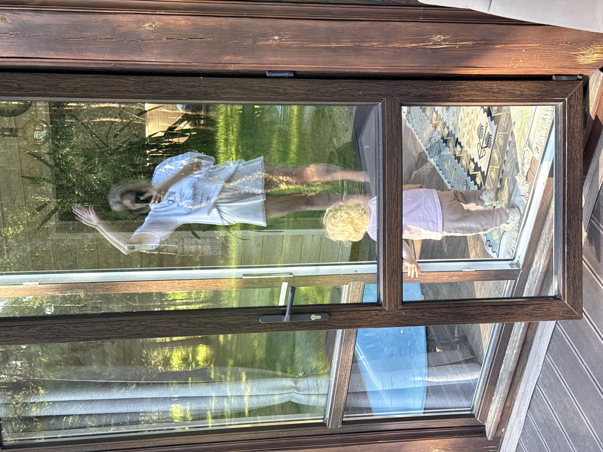 An eye-level shot captures a reflection in a large, dark brown-framed glass door or window on a sunny day in Копнино, Russia. The reflection features an adult woman and a young child, positioned indoors. The woman, with blonde hair, is wearing a white t-shirt with partially visible text that appears to read "BESTSELLER" and other words, and light-colored shorts. She is holding a smartphone, seemingly taking a photo or video of herself and the child. Her left arm is slightly raised. The child, with blonde curly hair and wearing a light pink top and beige pants, stands facing the woman, resting hands on the glass.
The transparent glass reveals a vibrant outdoor scene behind the reflections, including lush green grass, various trees, and possibly a wooden fence or structure in the background. The interior space, visible through the reflection, shows a patterned rug or carpet with geometric and floral designs, mostly in muted tones of brown, beige, blue, and black. A light fixture with an exposed filament bulb is also reflected in the upper left of the glass. Below the main reflections, a lower glass panel reflects blue-toned curtains or blinds from the interior. The robust wooden frame of the door/window includes a dark handle and lock mechanism. The exterior of the building features dark wooden paneling, suggesting a cabin or house setting. The bright lighting indicates it is daytime.