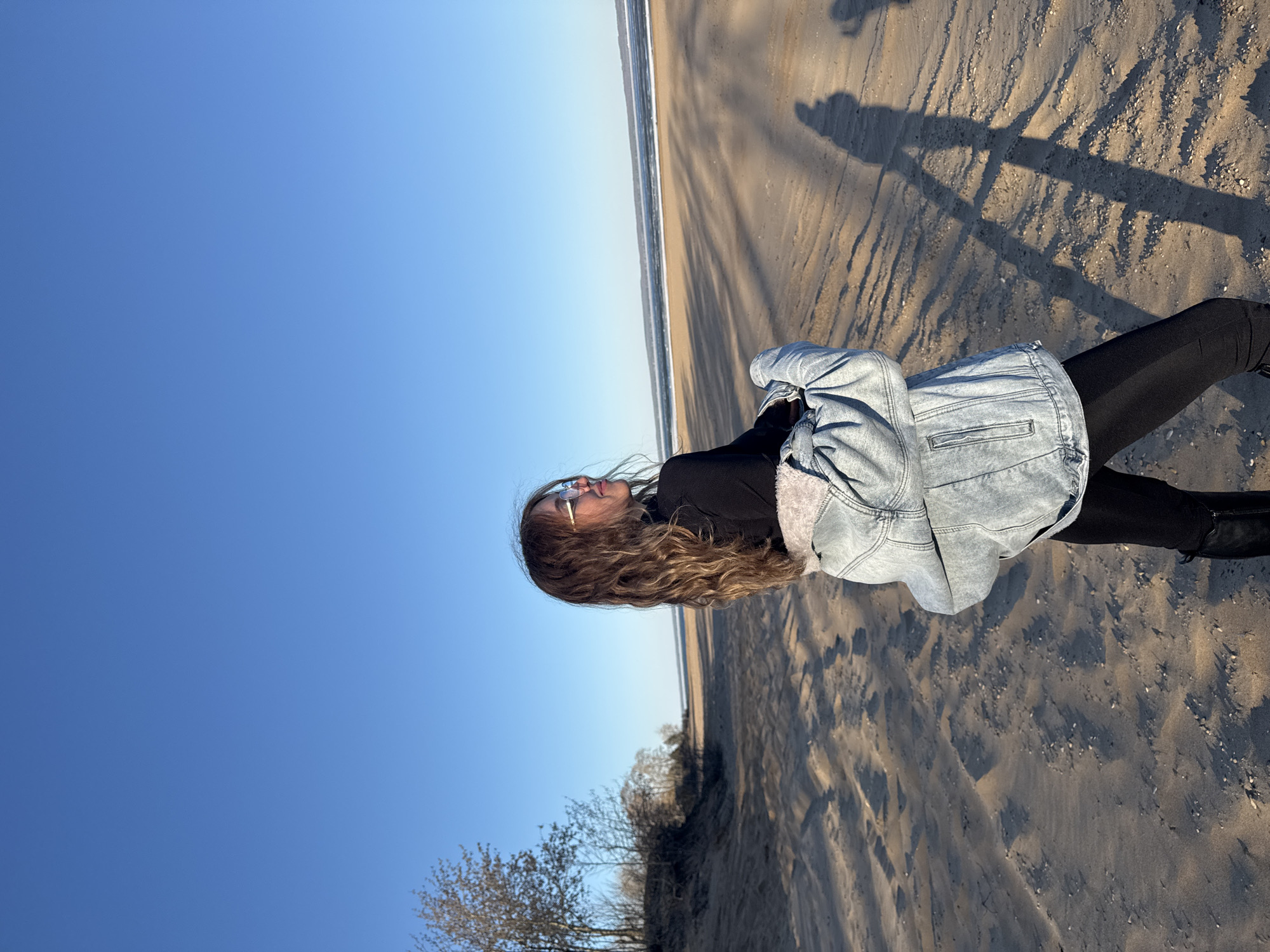 A young woman stands on a sandy beach under a clear, blue sky. She is wearing a denim jacket with a furry lining, a black top, and black leggings, and her long, wavy hair flows down her back. She is looking upwards, and the image is oriented so that the horizon line, with a thin strip of the sea, is horizontal. Her shadow stretches across the sand. In the lower left corner, there are bare trees. The location is likely near the city of Noviy, Russia, based on the prompt.