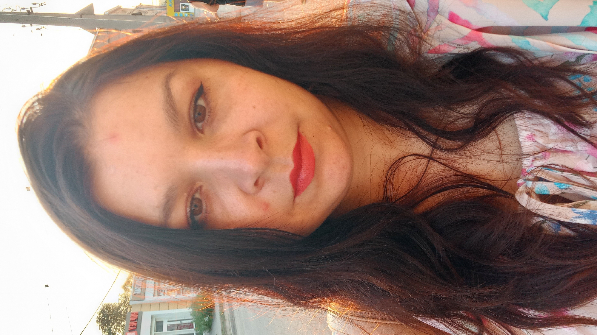 The image is a close-up selfie of a woman with dark, wavy hair and fair skin. She is wearing red lipstick and has eyeliner on. The background appears to be a street scene with buildings and a lamppost. The lighting suggests it is either sunrise or sunset. The woman is wearing a light-colored, patterned top. The scene is likely in Kropyvnytskyi, Ukraine, based on the style of the buildings and signage.
