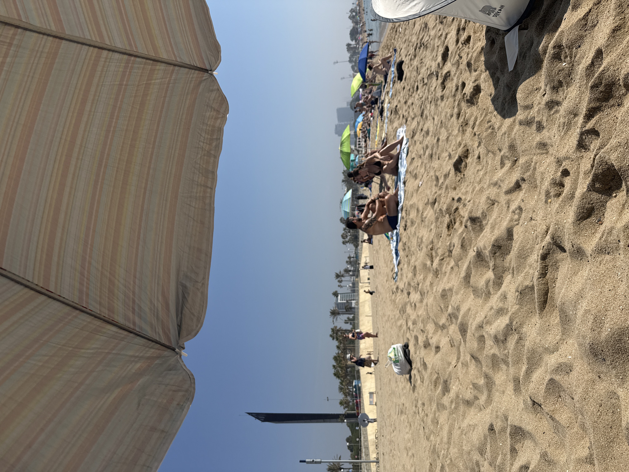 This is a sunny day at the beach in Barcelona, Spain. The foreground is dominated by sand, with ripples and footprints visible. A white plastic bag filled with items sits on the sand. In the middle ground, several people are scattered across the beach, some sitting under umbrellas, others walking. A couple is seated on towels, facing away from the camera. Behind them, more beachgoers are visible, along with colorful beach umbrellas and the Mediterranean Sea in the distance. In the background, palm trees line the promenade, and a distinctive tall, dark skyscraper stands out against the clear blue sky, likely the Torre Mapfre or Hotel Arts. The overall impression is a vibrant, active beach scene on a clear day.