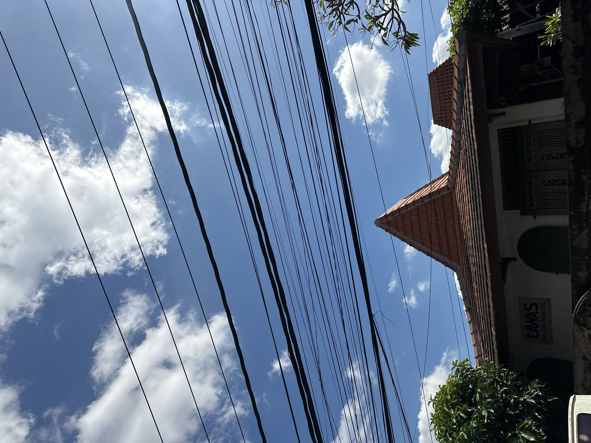 The image is a low-angle shot of a blue sky filled with scattered white clouds and an intricate network of black power lines. Below the sky, a portion of a building with a traditional tiled roof is visible. The building's facade is white with dark arched windows and a sign that reads "LAWAS OLD TEAK FURNITURE". To the left, a lush green tree is partially in frame, and to the right, branches of another tree with small white flowers are visible. The overall scene suggests an outdoor environment in a populated area, possibly a street in North Kuta, Indonesia, given the prevalence of such overhead wiring and architectural styles in the region. No people or animals are depicted in the image.