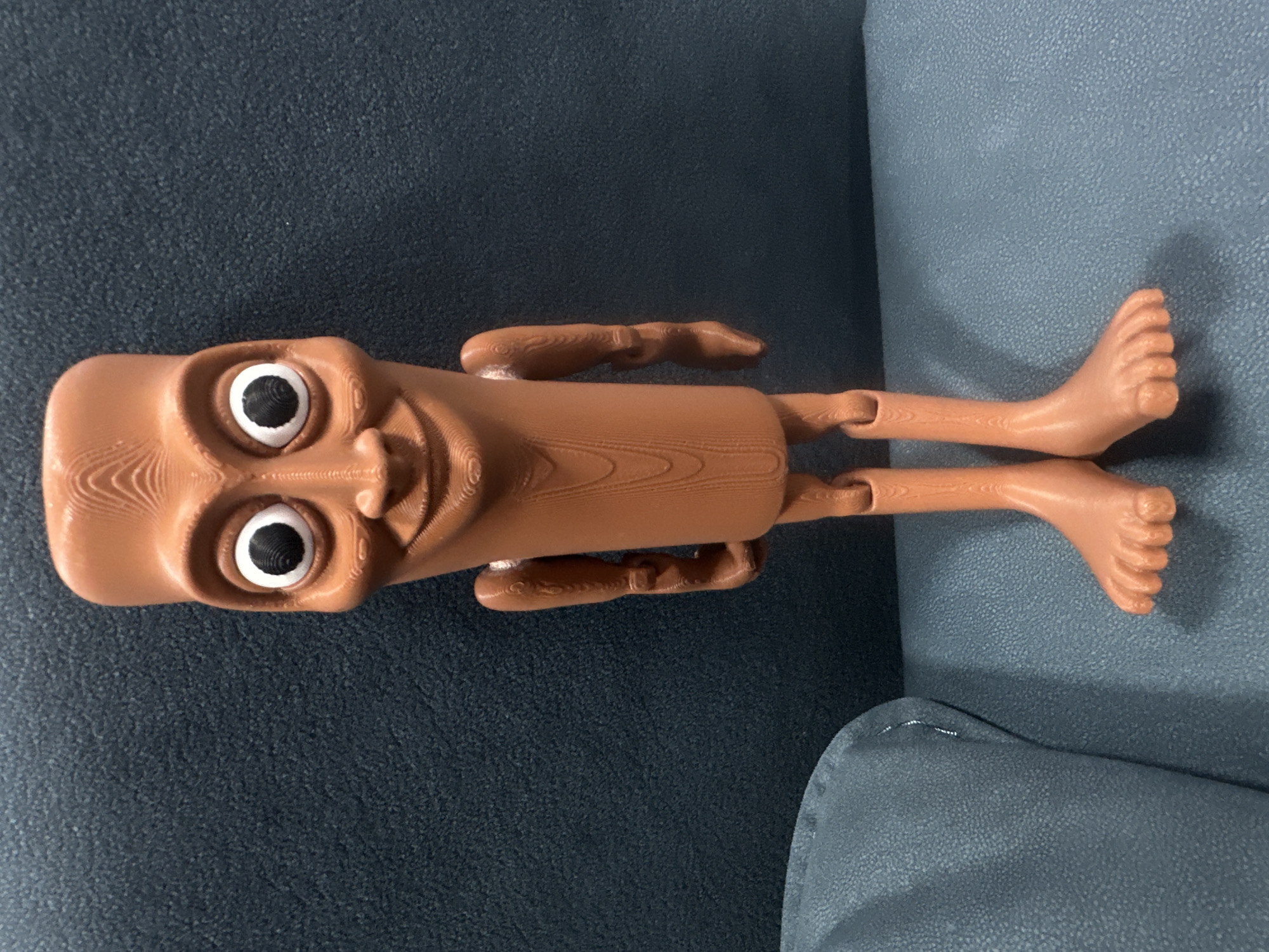 A brown, 3D-printed human figure lies on a textured dark blue and light blue/gray surface, possibly a sofa. The figure has a cylindrical body, exaggerated facial features including large eyes, and limbs with hands and feet. The environment does not provide any information to place the scene in Yerevan, Armenia.