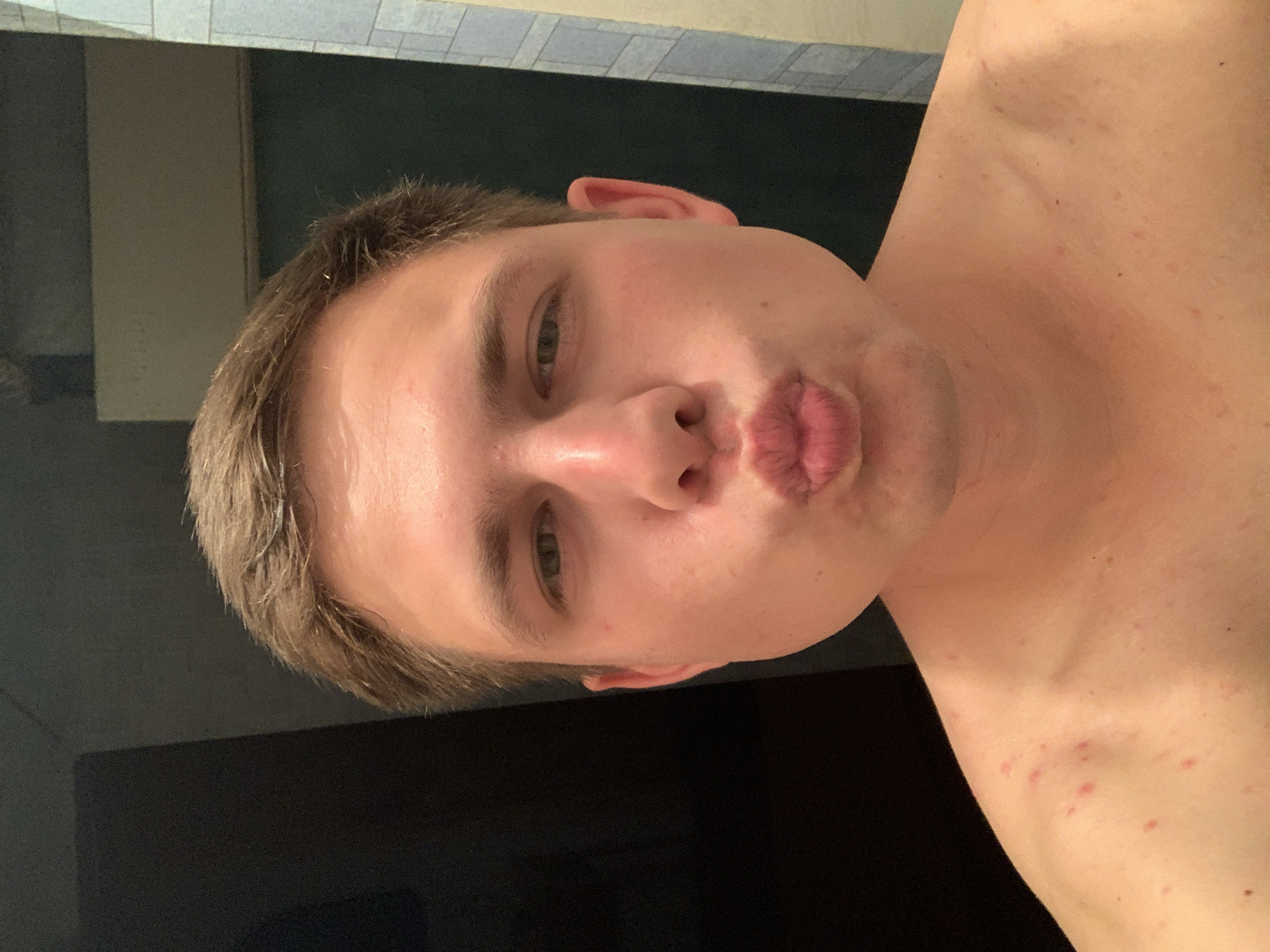 A close-up, head-and-shoulders selfie of a young, fair-skinned man with light brown hair. He is looking directly at the camera with his lips puckered as if for a kiss. His skin is slightly red and appears to have some minor blemishes on his shoulders. The background is dark and out of focus, with a hint of a tiled wall on the right side of the frame. The overall lighting is warm and appears to be coming from the front. The man is shirtless.