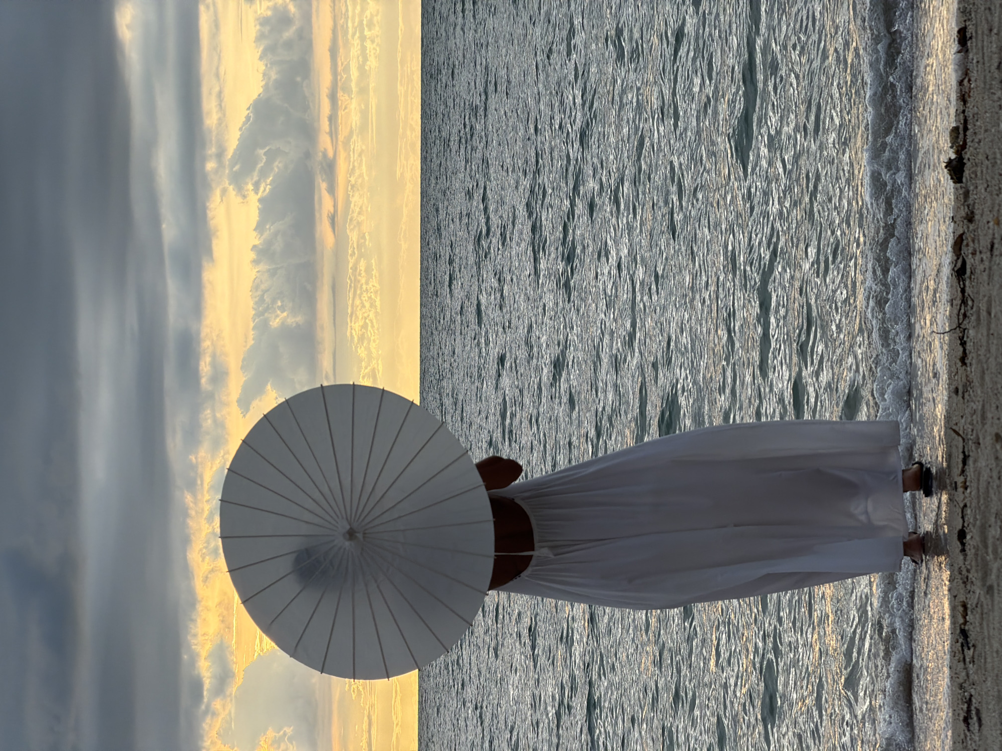 A person stands on a sandy shore, holding a white parasol. They are wearing a long, flowing white dress that reaches to their ankles. The person is facing away from the camera, towards the ocean. The water is calm and reflects the light of the setting sun. The sky is filled with dramatic clouds, tinged with orange and yellow from the sunset. The overall mood is serene and peaceful. It appears to be late afternoon or early evening. The location is a beach in Baan Plai Laem, Thailand.