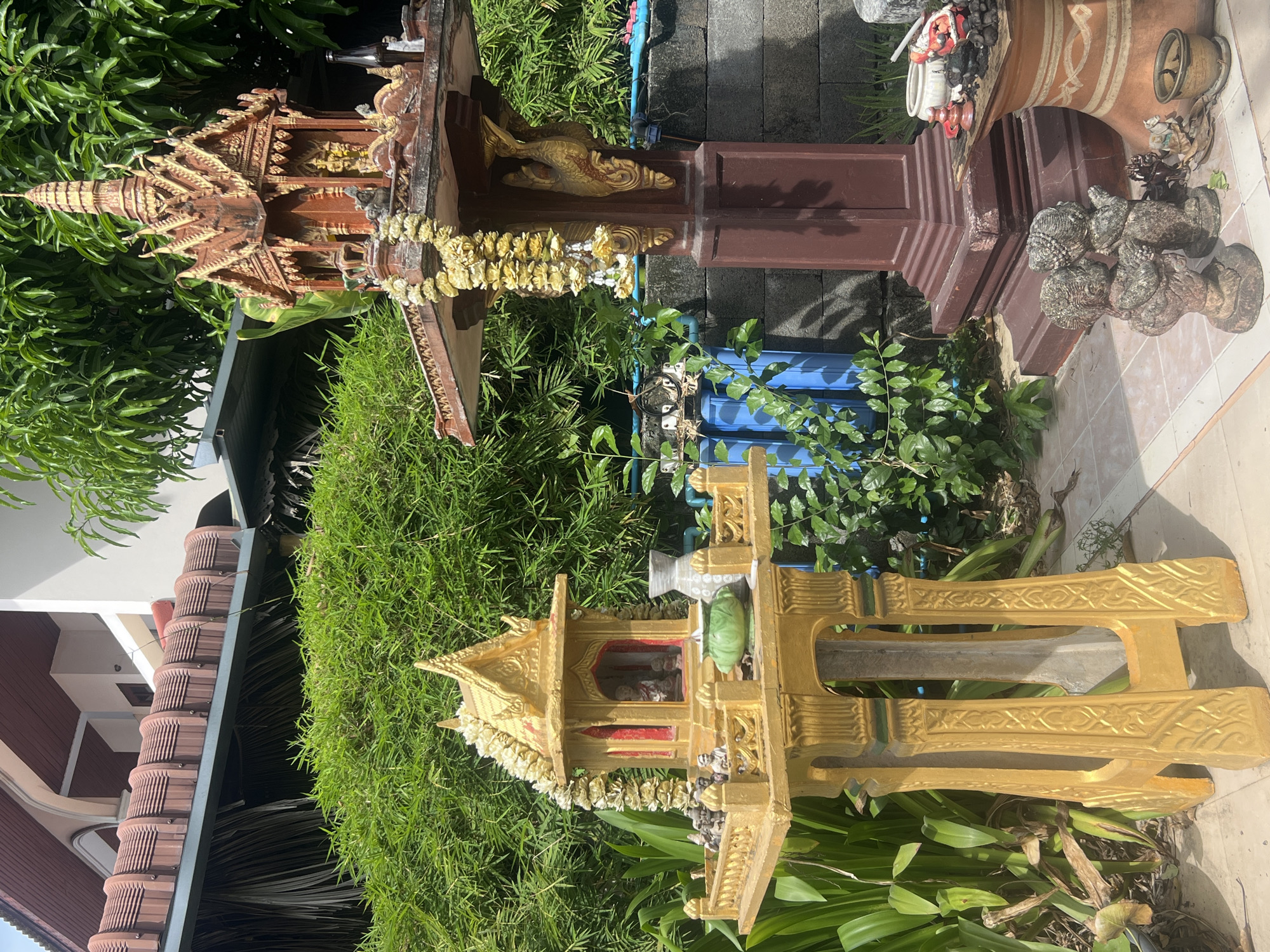 Two spirit houses stand in a garden setting in Baan Lamai, Thailand. One is a brown wooden structure with intricate carvings and a multi-tiered roof. The second is a golden-colored house with a simpler, yet ornate design. Both are adorned with garlands of flowers. They are set amongst green vegetation, including plants and grass. In the background, a building with a terracotta-tiled roof is partially visible. The houses are on a tiled surface. There is a brown pedestal near the brown house. There are also blue water tanks and some other decorative items next to the brown house.