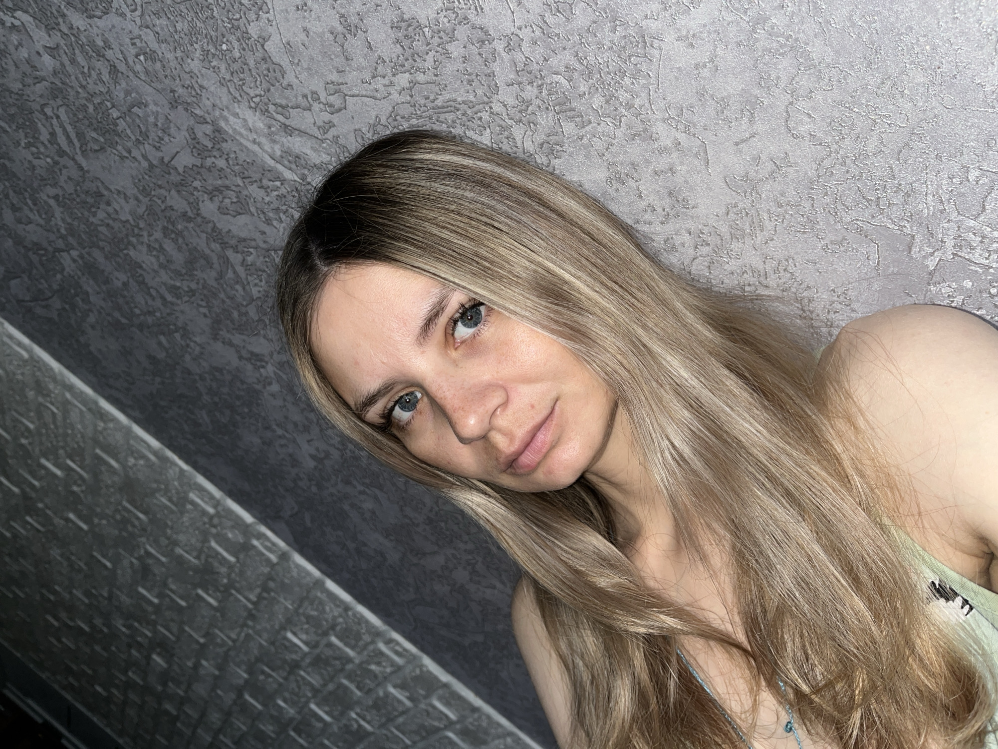 A young woman with long, blonde hair and blue eyes looks directly at the camera. She is wearing a light green top with a small, embroidered design on it, and a thin, blue necklace. The background consists of a textured, gray wall and a section of what appears to be a gray, brick-like wall. The image does not provide enough information to identify a specific location in Chaikovsky, Russia.