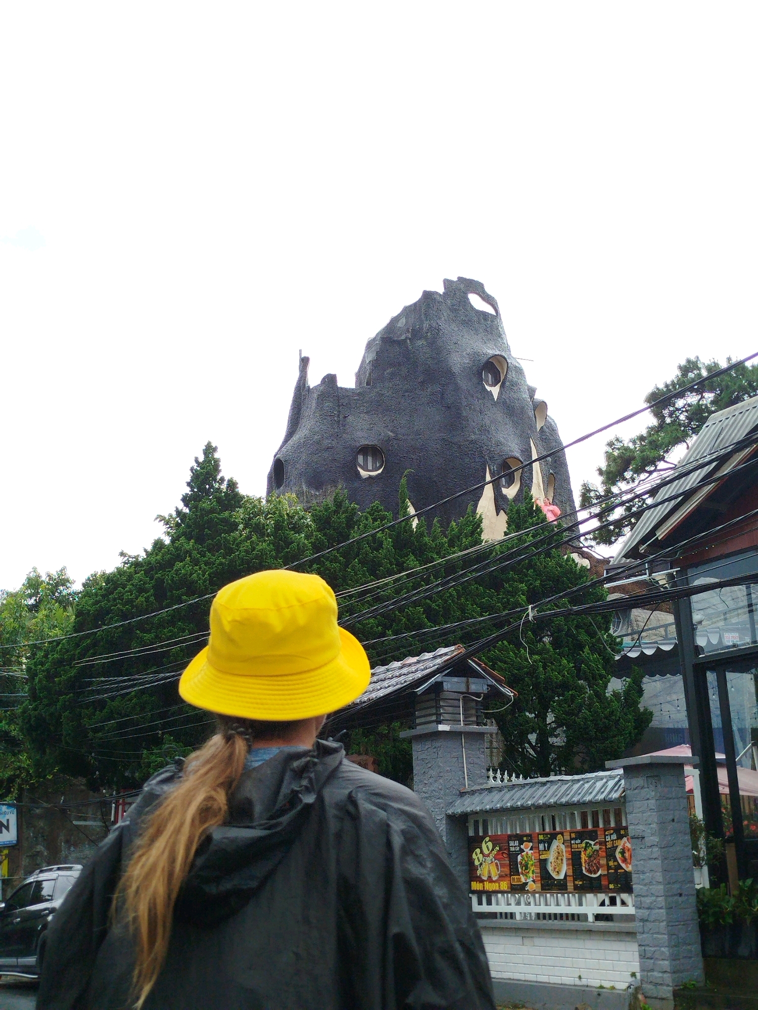 In Da Lat, Vietnam, a person wearing a yellow bucket hat and a dark rain jacket faces away from the camera, their blonde hair pulled into a ponytail. They stand in front of a distinct, dark-colored building with an unusual, organic shape, featuring multiple irregularly shaped windows and a texture resembling rock or bark. The building is partially obscured by lush green trees. To the right of the main building, a smaller structure with a corrugated metal roof is visible, and further to the right, a glass-fronted building with a dark frame is present. In the foreground, a grey stone fence with a white railing displays several posters advertising food. The sky is overcast and white, suggesting cloudy weather. Power lines crisscross the scene, indicating an urban or semi-urban setting.