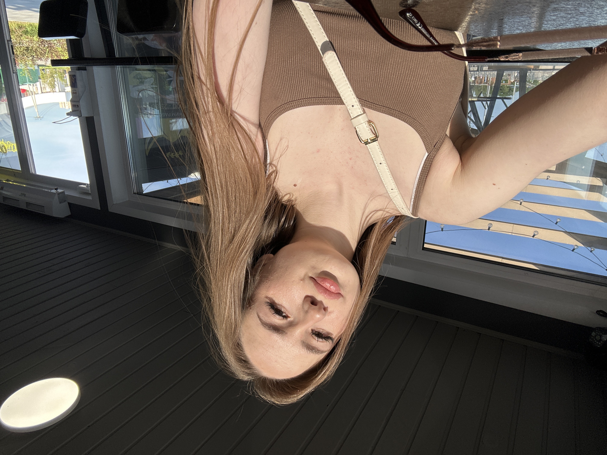 An upside-down, eye-level selfie shows a young woman indoors next to large windows on a bright, sunny day. The woman has long, straight light brown hair that frames her face. She looks directly at the camera with a calm expression, her lips slightly parted. She is wearing a brown ribbed tank top, and a cream-colored cross-body bag with a gold buckle strap is visible across her chest. A pair of sunglasses with dark red frames is hooked onto her top, and the text "UV PROTECTED" and "CAT.3 CE" is visible on one arm.
The setting appears to be an interior space, possibly a cafe or restaurant, characterized by dark, horizontally ribbed flooring. A prominent oval-shaped white light is reflected or embedded on the floor in the lower left. Large windows occupy the background, showcasing a clear blue sky and brightly lit outdoor scene with reflections of buildings or structures. A white heater or vent unit is installed below the leftmost window. Between two window panes, a wall-mounted device, possibly a hand sanitizer dispenser or payment terminal, is visible. String lights are strung across the outdoor view through the windows. Partial text "МРАВ" and "00-23:00" can be seen in a reflection on one of the windowpanes. Strong sunlight casts distinct shadows and highlights throughout the scene.