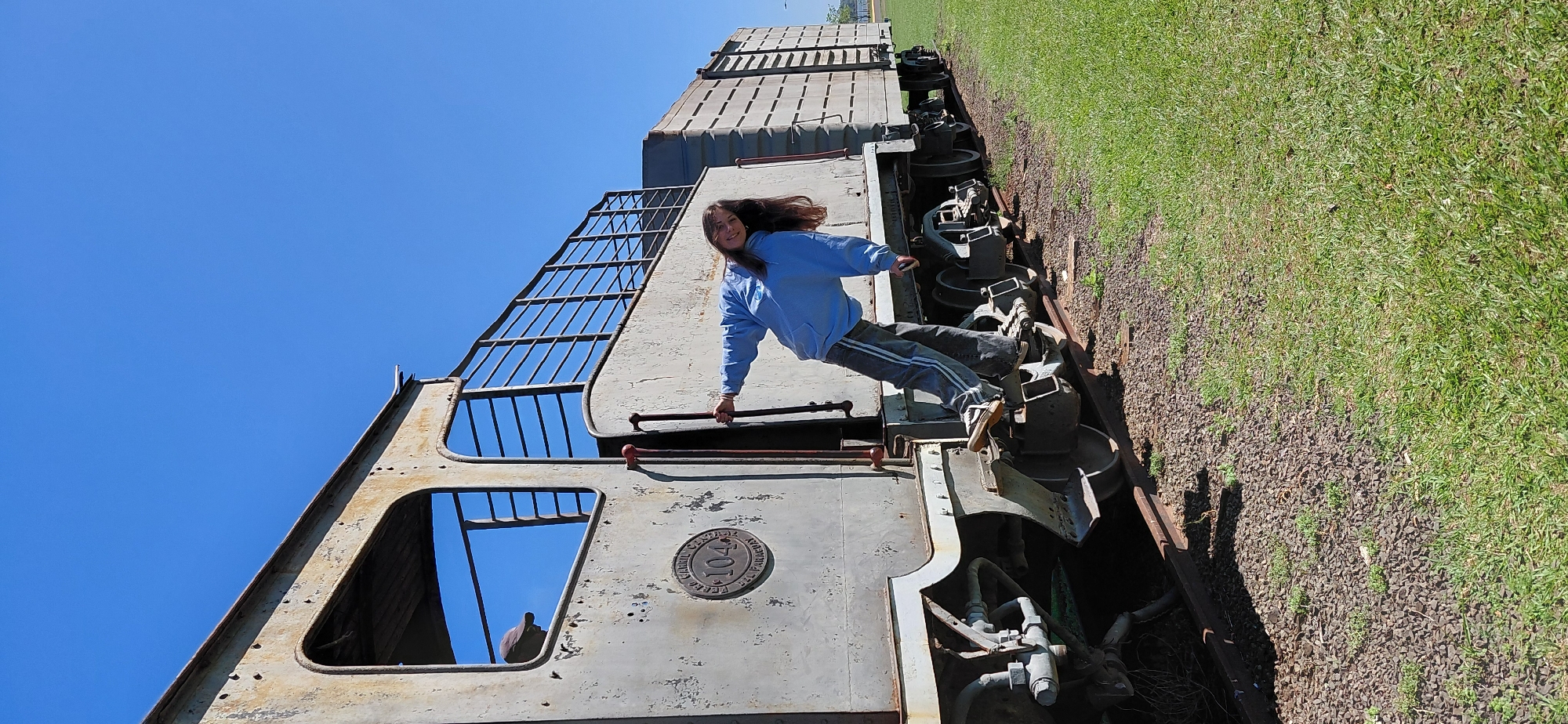 A young person wearing a light blue hoodie and dark pants with white stripes is posing on the side of an old, rusty train car. They are leaning back, with their right hand gripping a vertical metal bar and their left hand outstretched. Their feet are placed on the train's mechanism and wheel assembly. The train is on a track next to a grassy area under a clear blue sky. In the background, more train cars and a distant view of water can be seen. A person wearing a dark cap is visible through the window of the train car. A circular metal plaque with the number "104" and text is affixed to the side of the train car. This train is located at the Train Museum in Encarnacion, Paraguay.