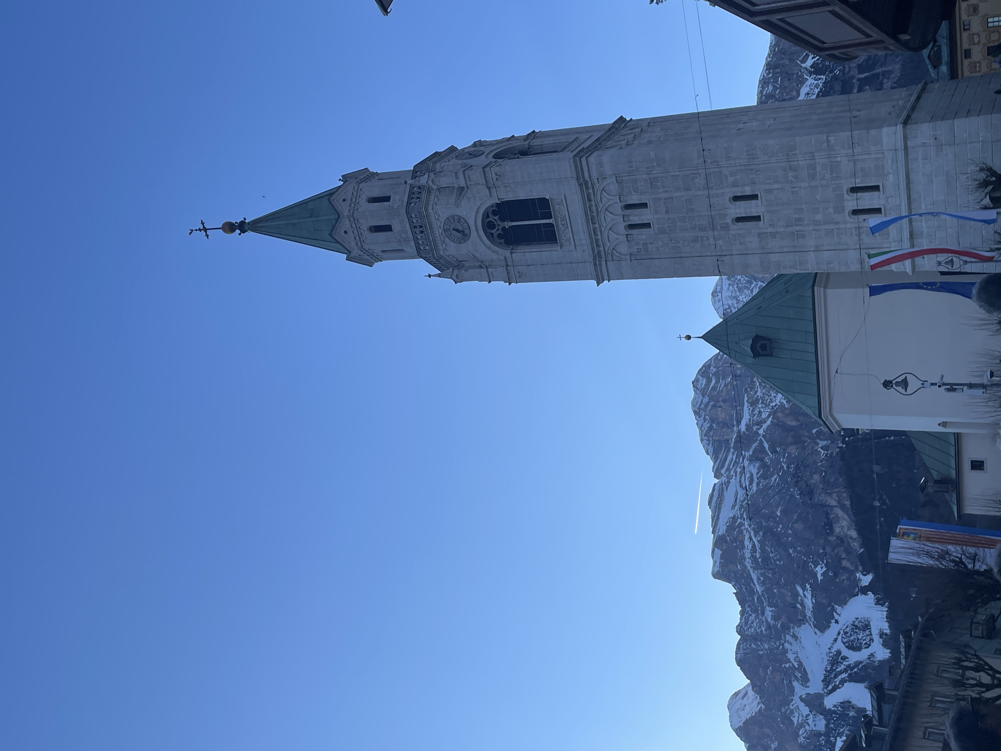 A low-angle shot showcases the bell tower of the Basilica Minore dei Santi Filippo e Giacomo in Cortina d'Ampezzo, Italy. The tower's intricate architectural details are visible, contrasted against a clear blue sky. Nearby, a building with a green roof and a white facade stands next to the tower. In the background, snow-covered mountains rise behind the buildings. Italian and EU flags fly at the base of the tower. A faint contrail is visible in the sky behind the mountain. The shot appears to be taken during daylight hours.