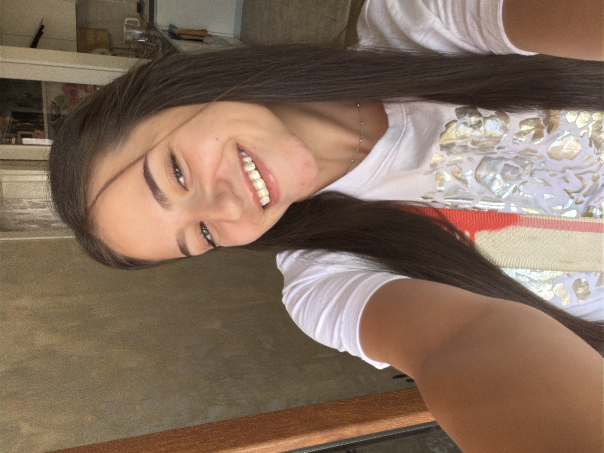 A young woman with long, dark brown hair is taking a selfie indoors. She is smiling and wearing a white t-shirt with a floral pattern. The background is blurred but appears to be a domestic setting, possibly a kitchen with cabinets and a countertop visible. There is a wooden structure on the left side of the image. The image quality is somewhat grainy.