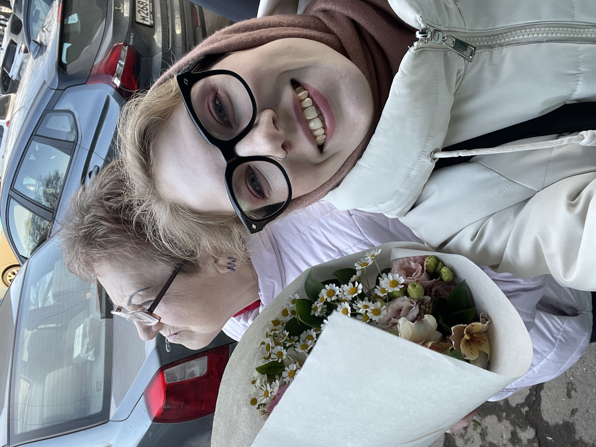 Two people, likely a young woman and an older woman, are posing for a picture while holding a bouquet of flowers. The younger woman is wearing large, black-framed glasses and a light-colored hooded jacket. The older woman is wearing glasses with a thinner frame. In the background, there are parked cars, including a grey one with the license plate "M268K." Based on the license plate, the location is the city of Izhevsk, Russia. The bouquet contains a mix of small white daisies and other flowers. The bouquet is wrapped in white paper. The scene is outdoors and appears to be on a city street.