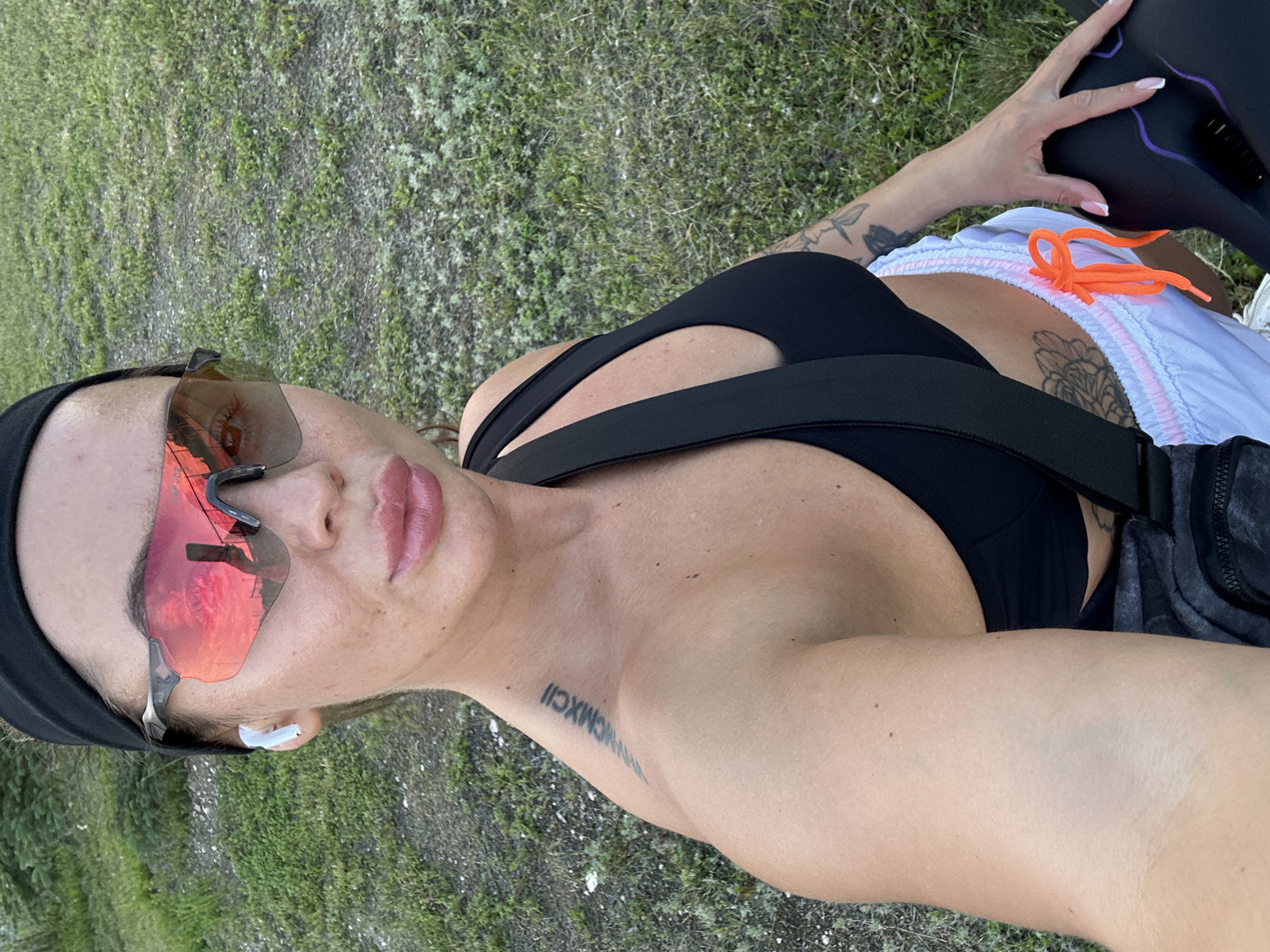 A close-up selfie shows a woman wearing a black headband, sunglasses with red lenses, and a black sports bra. She has a tattoo on her left shoulder blade. She is also wearing white shorts with pink trim and an orange drawstring. The background is a grassy area with some gravel. The woman is outdoors, possibly in a park or recreational area.