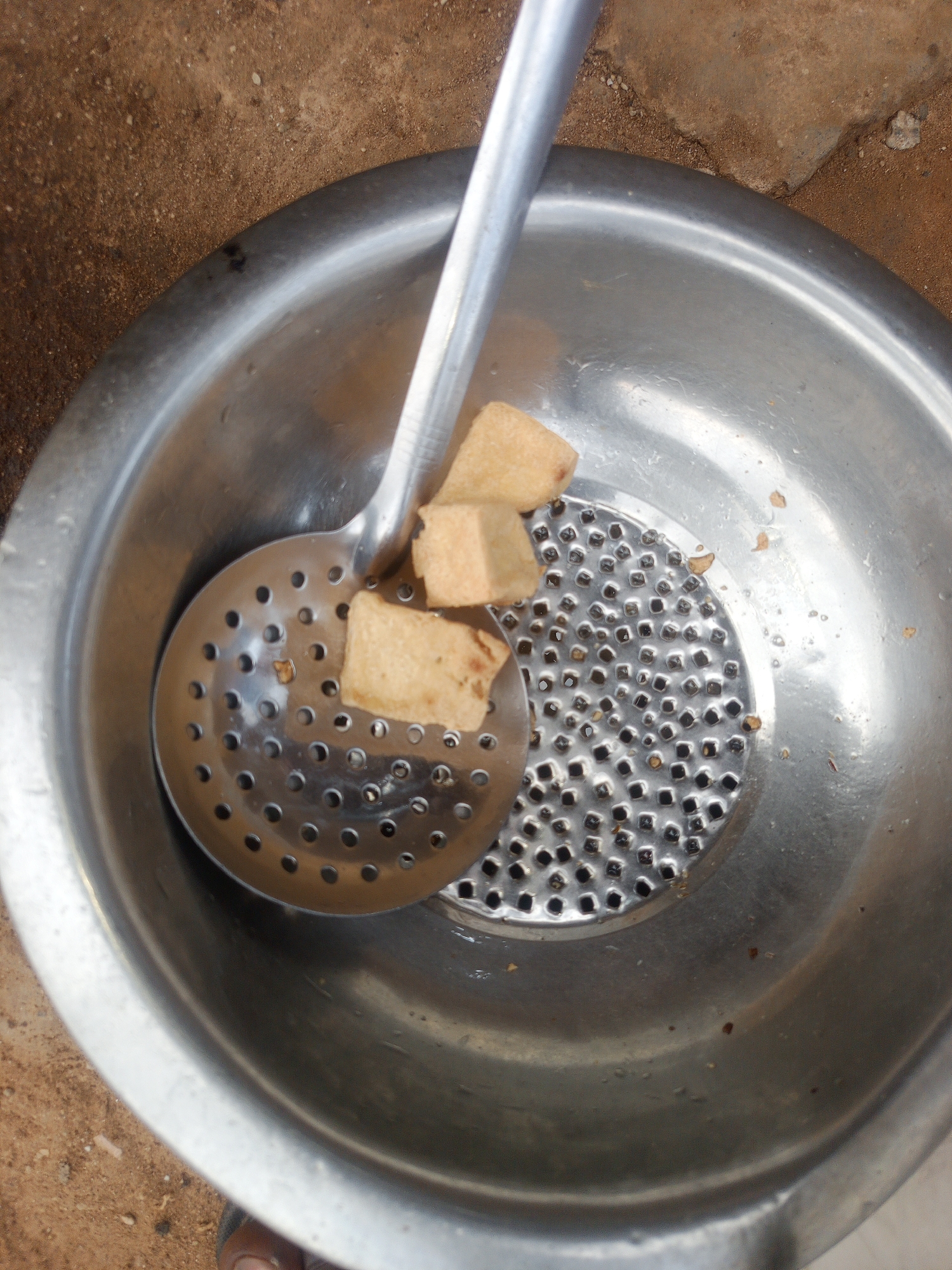 A close-up, high-angle shot shows a metal colander inside a larger metal bowl. The colander has many small, square-shaped holes. A metal slotted spoon rests on top of the colander. Three pieces of fried food, appearing to be cubes of fried yam or plantain, are on the slotted spoon. The food is golden brown. The background is a coarse, brown, earthy surface. The scene is likely outdoors, given the natural texture of the ground. The image is in focus, with good lighting. There are no people or animals clearly visible. The overall impression is one of food preparation or draining. The setting, with the earthy ground, is consistent with an outdoor environment in Abuja, Nigeria.