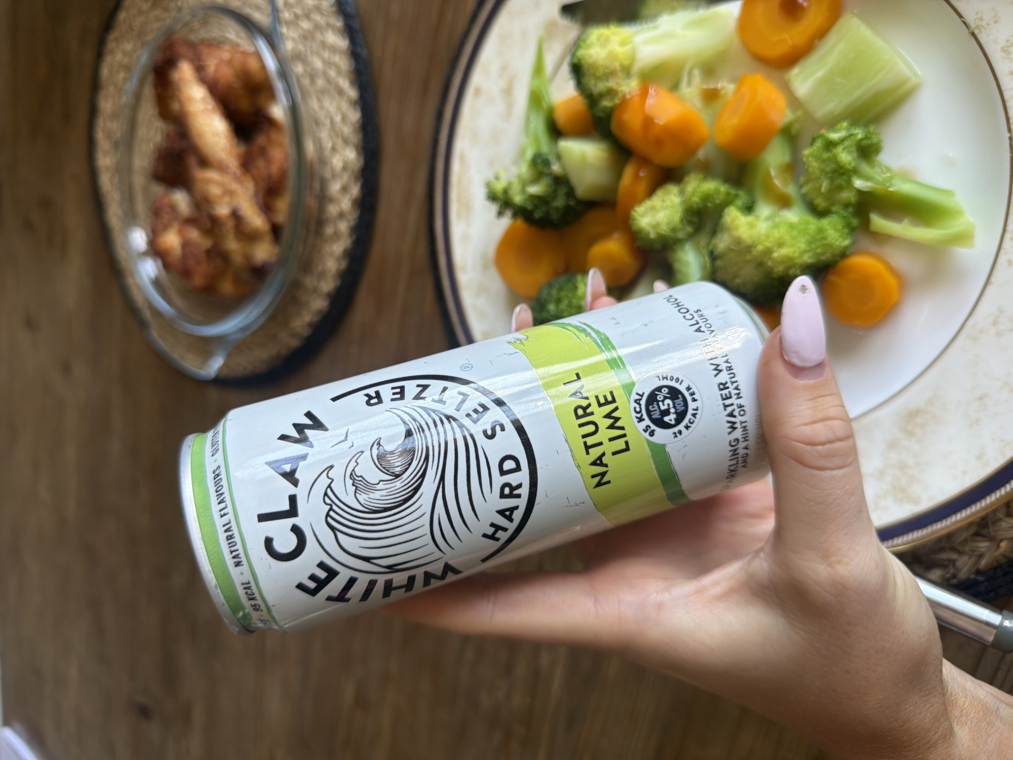 A close-up, high-angle shot shows a hand holding a White Claw Natural Lime Hard Seltzer can in front of a plate of steamed vegetables and a glass bowl of chicken. The can is white with black lettering and a green label that reads "NATURAL LIME." The vegetables on the plate include broccoli, carrots, and celery. The chicken in the bowl appears to be roasted or fried. The background is a wooden table. The scene is indoors, likely in a home or restaurant. The lighting is natural, coming from a window or overhead source.