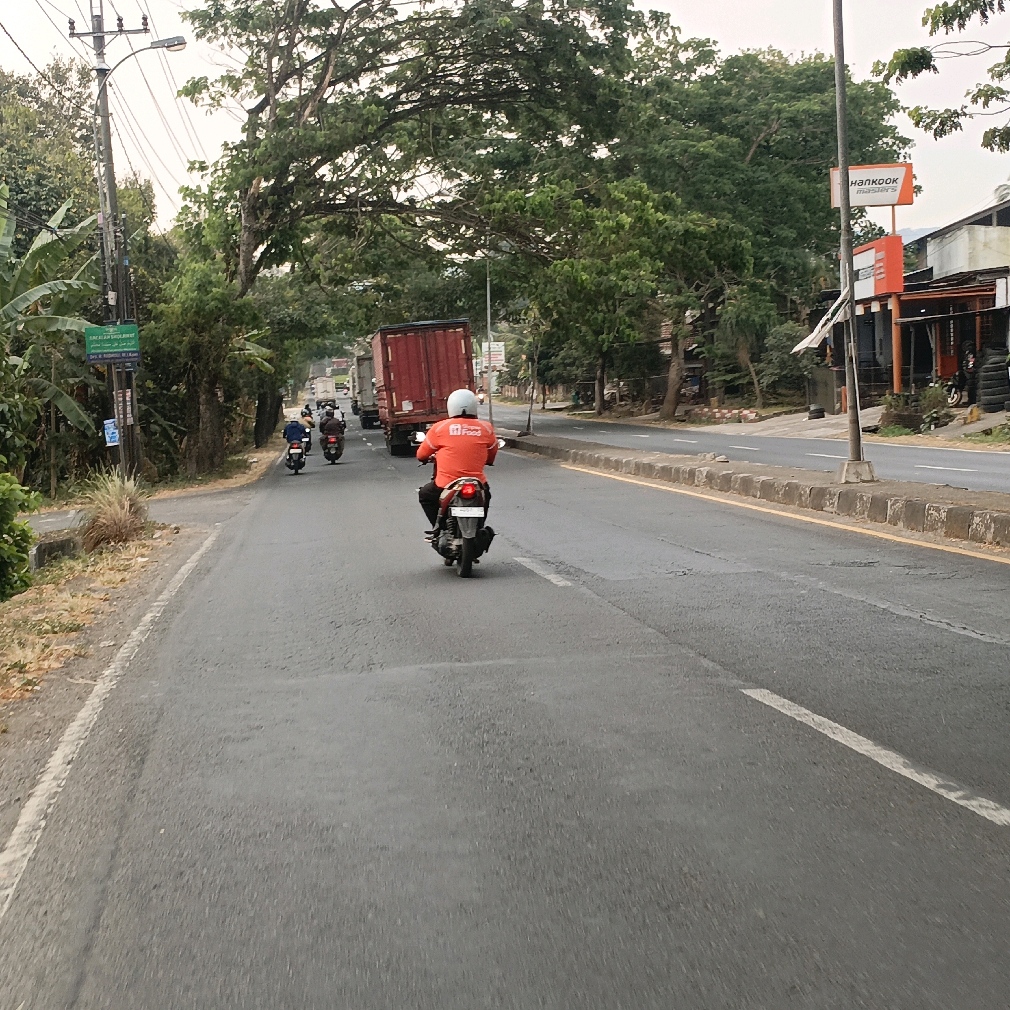 In Salatiga, Indonesia, a motorcycle rider wearing a bright orange jacket and white helmet is traveling down a paved road. The rider is positioned in the center of the lane, with a red cargo truck in front and other motorcycles behind. The road has a white line marking the lane and a yellow line along the curb. On the left side of the road, there are trees, a utility pole with a green sign, and some foliage. On the right side of the road, there is a building with an orange and white sign that says "HANKOOK masters". The sky is overcast.