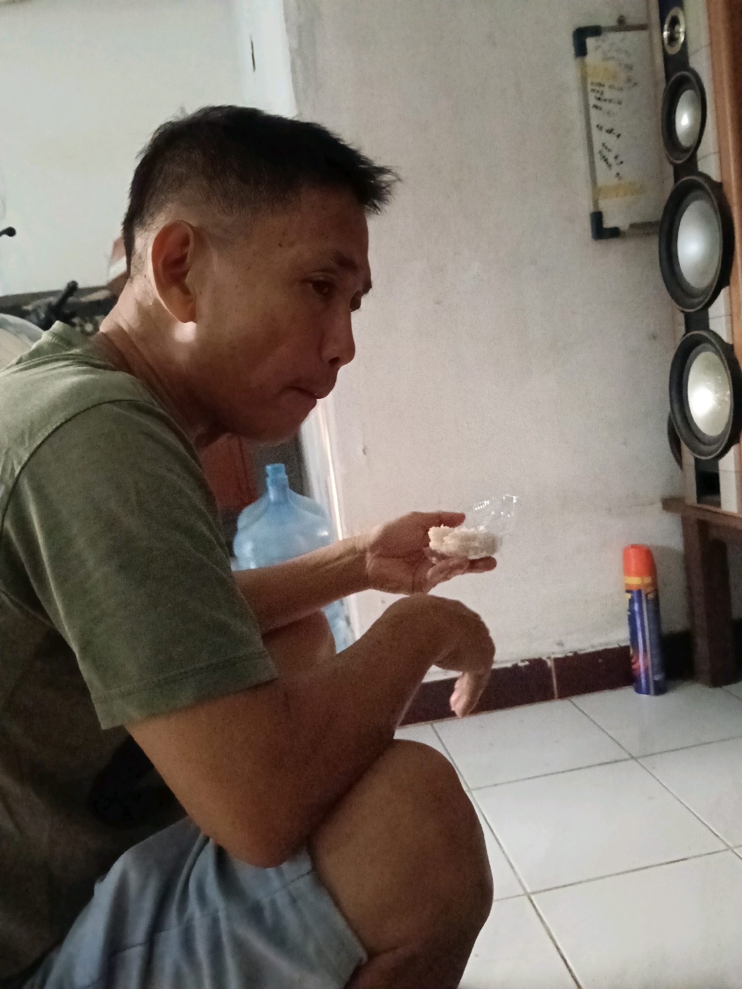 The image shows an Asian male with short hair, wearing a green t-shirt and blue shorts, sitting indoors. He is holding a small clear plastic container filled with a white substance, possibly food, and appears to be eating or about to eat from it. The background includes a wall with speakers mounted vertically, a whiteboard with writing, a large blue water jug, and a spray can on the floor. The lighting suggests an indoor environment, possibly a home or a simple living space. The flooring appears to be tiled. The location is identified as Jatimulya, Indonesia.