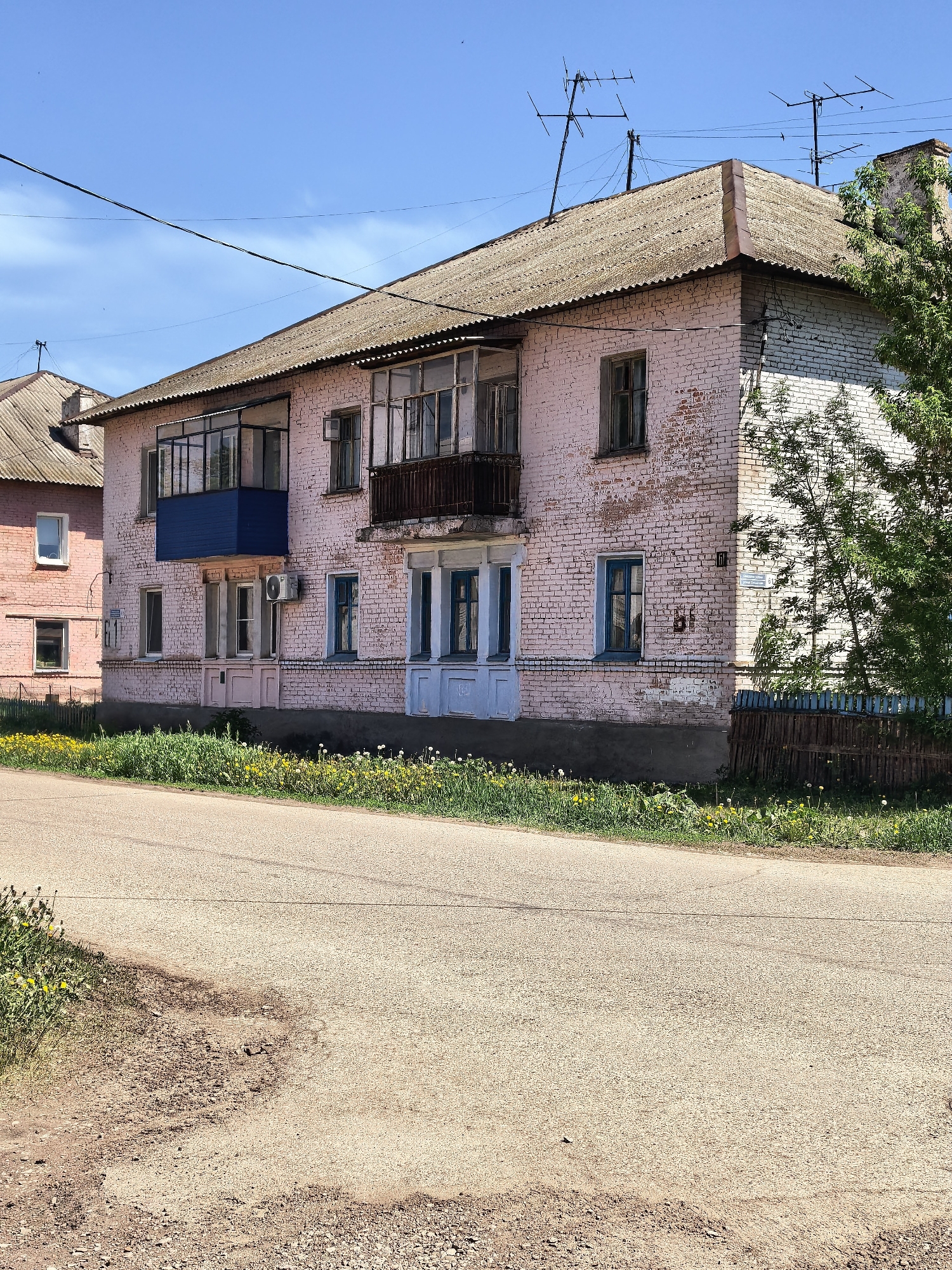 The image shows a two-story pink brick building with blue accents in Sterlitamak, Russia. The building has a dark gray corrugated roof with multiple antennas on top. Balconies with metal railings are attached to the second floor. The building appears aged and slightly dilapidated. A smaller, similar pink building is visible in the background. A paved road runs in front of the building, with grass and dandelions growing alongside it. A partial wooden fence is near the corner of the building, partially obscured by a tree. Power lines run across the sky. The weather is sunny with a clear blue sky.