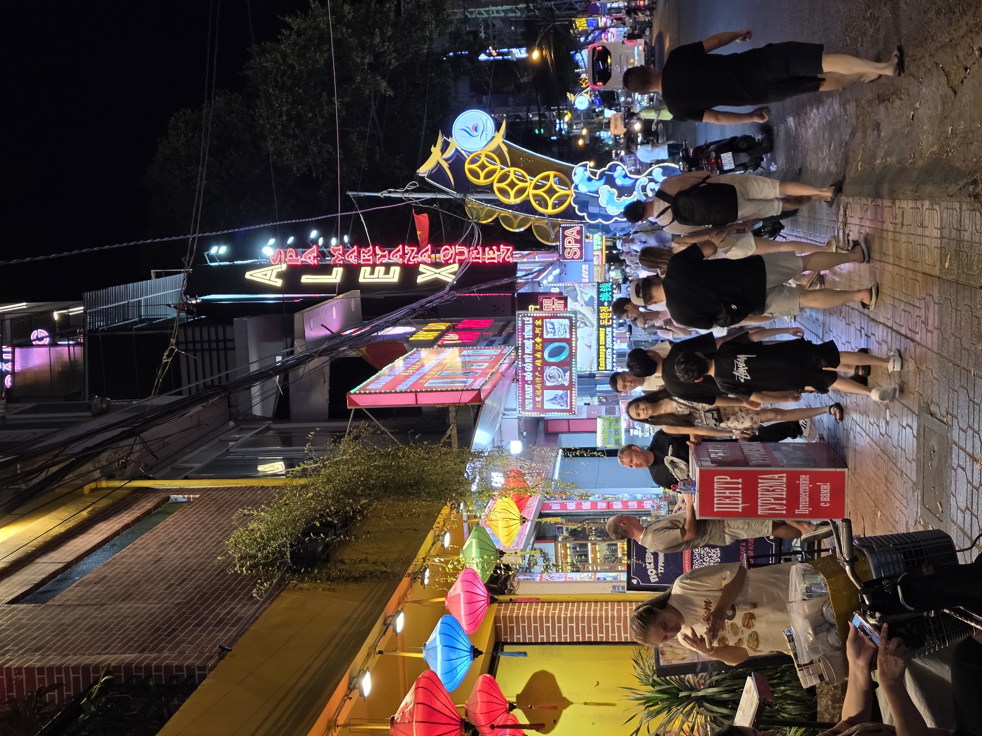This image depicts a vibrant and bustling street scene at night in Nha Trang, Vietnam. The setting is a commercial thoroughfare lined with various shops and establishments, all brightly illuminated by numerous neon signs and artificial lights. Overhead power lines crisscross the dark sky, characteristic of urban areas in Southeast Asia.
Many people are visible, walking along the sidewalks and gathered in groups. Most are seen from behind or in profile, suggesting movement and an active environment. One woman is observing items on a stall, while others appear to be browsing or conversing. The atmosphere seems lively and recreational, indicative of an evening out.
Notable details include a profusion of colorful neon signage advertising businesses, often in multiple languages such as Vietnamese, English, Russian, Korean, and Chinese, signaling a popular tourist destination. Prominent signs include "ALEX," "SPA," and a circular sign displaying "NHA TRANG." A red stand prominently features "ЦЕНТР ТУРИЗМА" (Center of Tourism) and "Путешествуйте с нами!" (Travel with us!) in Russian. Other signs mention "MINI MART - ĐỒ GỖ MỸ NGHỆ TỪNG LÝ" (Mini Mart - Tuong Ly Fine Art Wood), "Exchange money," and "FRENCH PANDANES" with "Французские Блины" (French Pancakes). Brightly colored lanterns in pink, blue, and yellow are strung above a section of the sidewalk, enhancing the festive night ambiance. The ground is a mix of pavement and cobblestones, and some greenery is visible from planters.