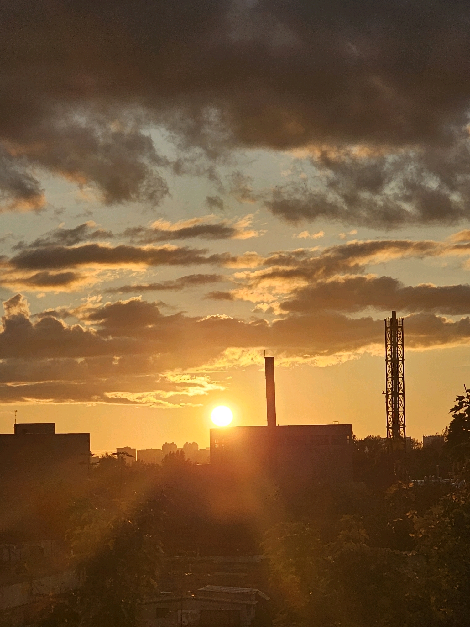 The image depicts an industrial cityscape at sunset in Khimki, Russia. The sky is filled with dramatic, golden and grey clouds. In the foreground, silhouetted buildings and trees obscure the ground level. Prominently featured are two industrial structures: a tall, slender tower with a latticed framework on the right side of the image, and a shorter, rectangular building with a smokestack on the left. The sun, a bright orb, is setting just above the horizon, casting a warm, orange glow over the scene and creating lens flare. The overall impression is one of an industrial area bathed in the dramatic light of a setting sun.