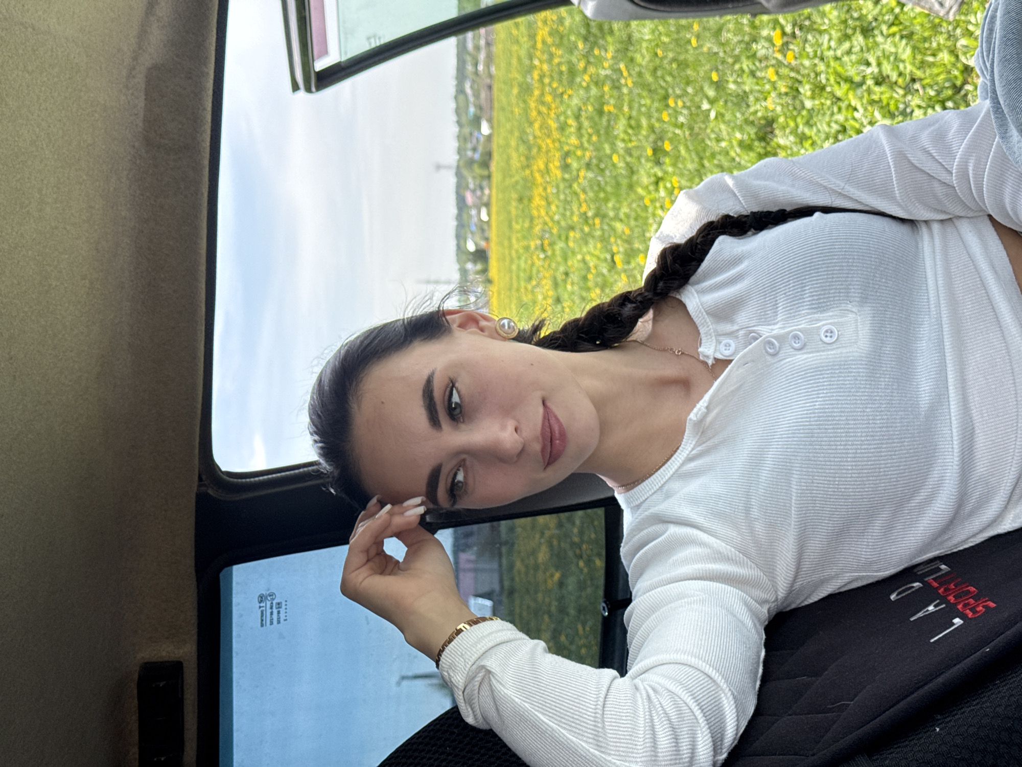 A woman is sitting inside a vehicle, possibly a car, with a landscape visible through the open door. She is fair-skinned with dark hair styled in a braid. She is wearing a white, long-sleeved, button-down shirt and a gold bracelet on her left wrist. She is looking towards the camera with a slight smile and her left hand is near her face. Outside the vehicle, there is a field of yellow flowers and some buildings in the distance. The location is Фоки, Russia.